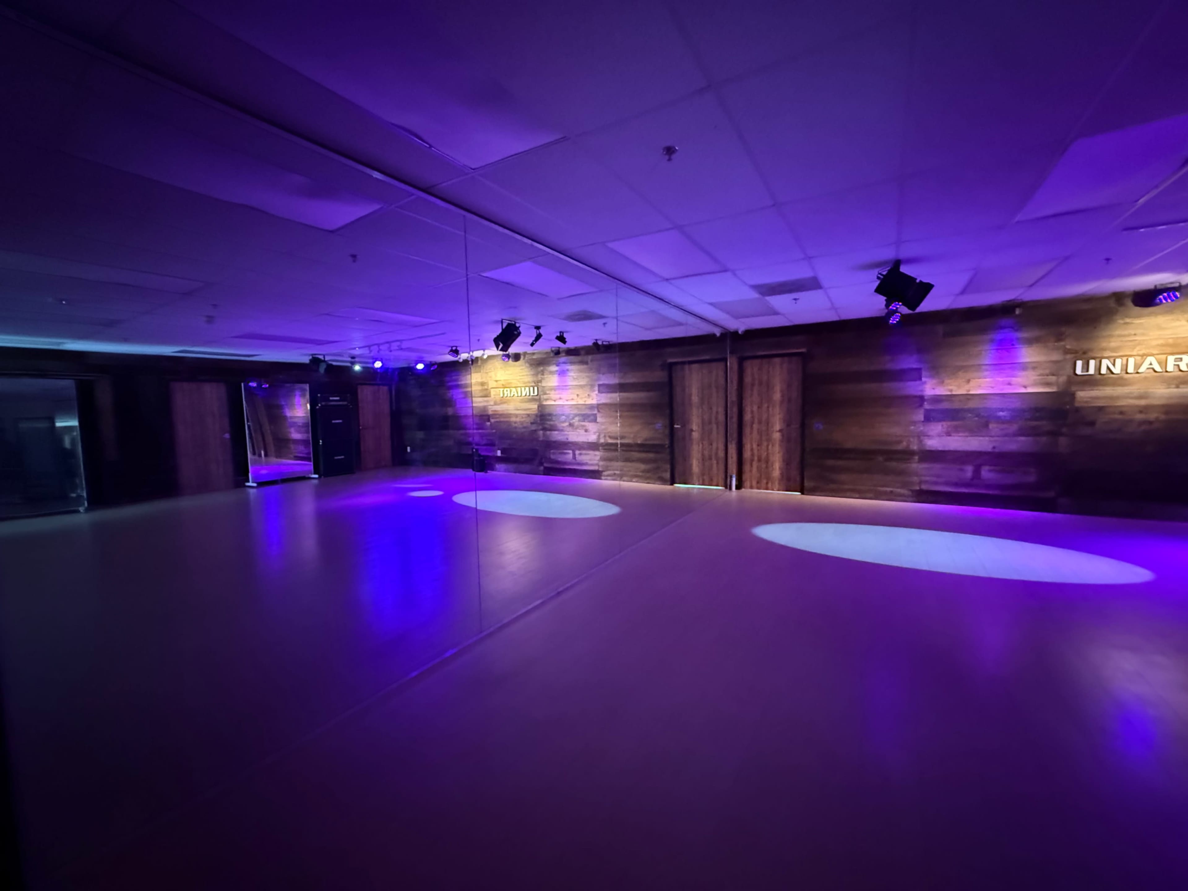 The image shows an empty dance studio with wooden walls, a mirrored wall, and purple lighting.