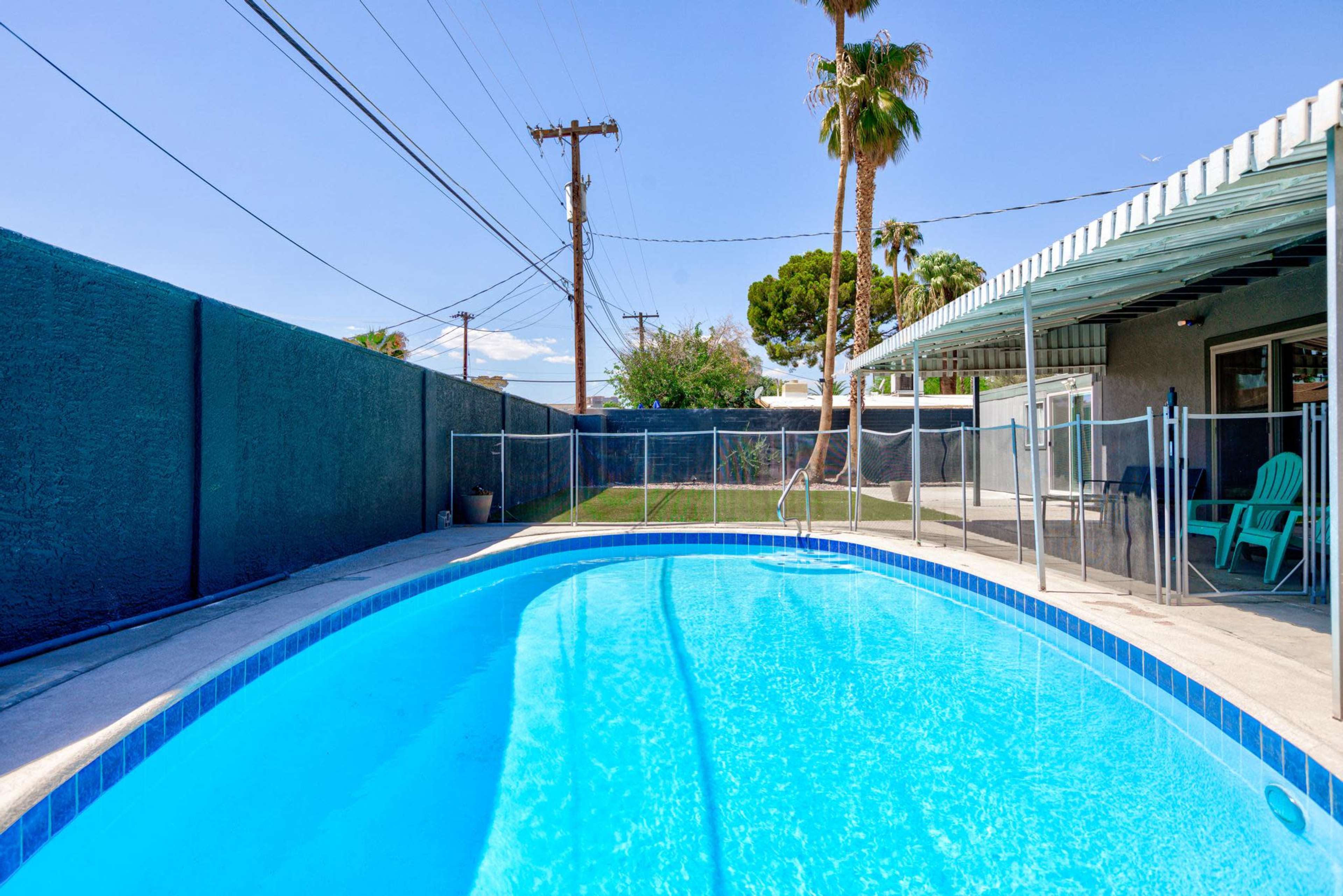 A clear blue swimming pool is located in a fenced backyard with palm trees and utility poles visible in the background.
