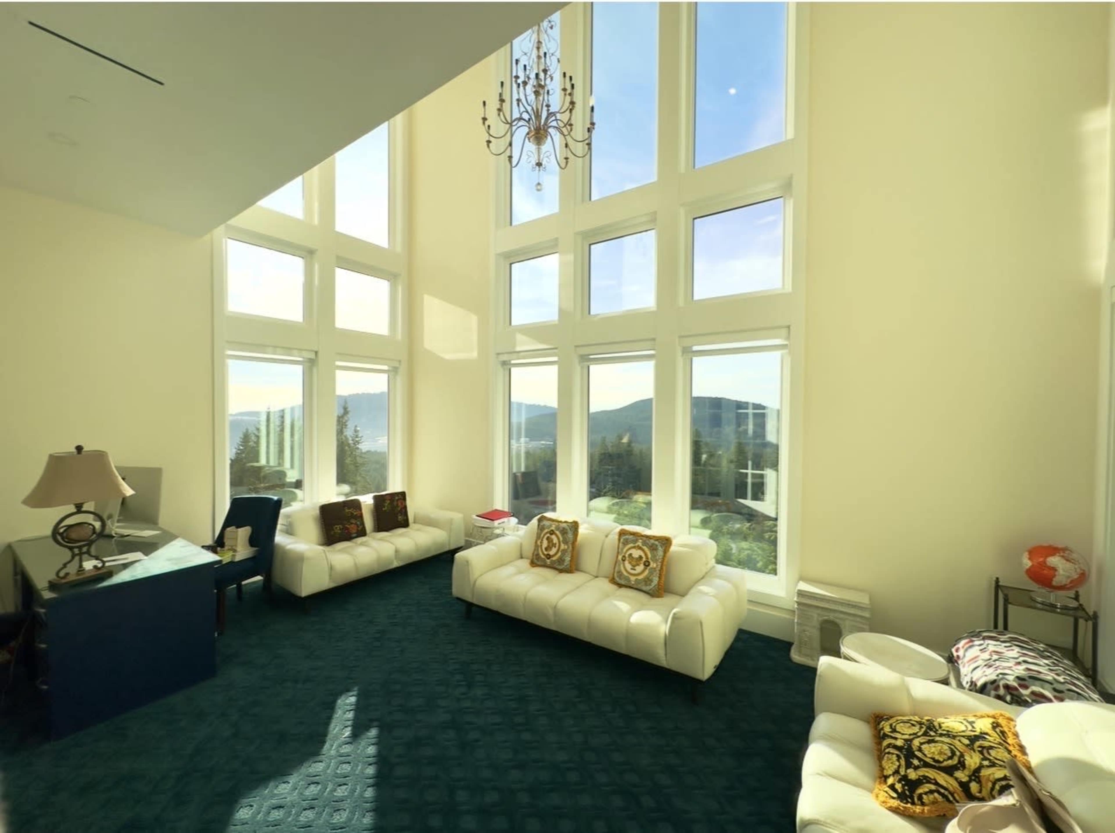 The image shows a spacious living room with large windows, two white leather sofas, a green carpet, and a chandelier.