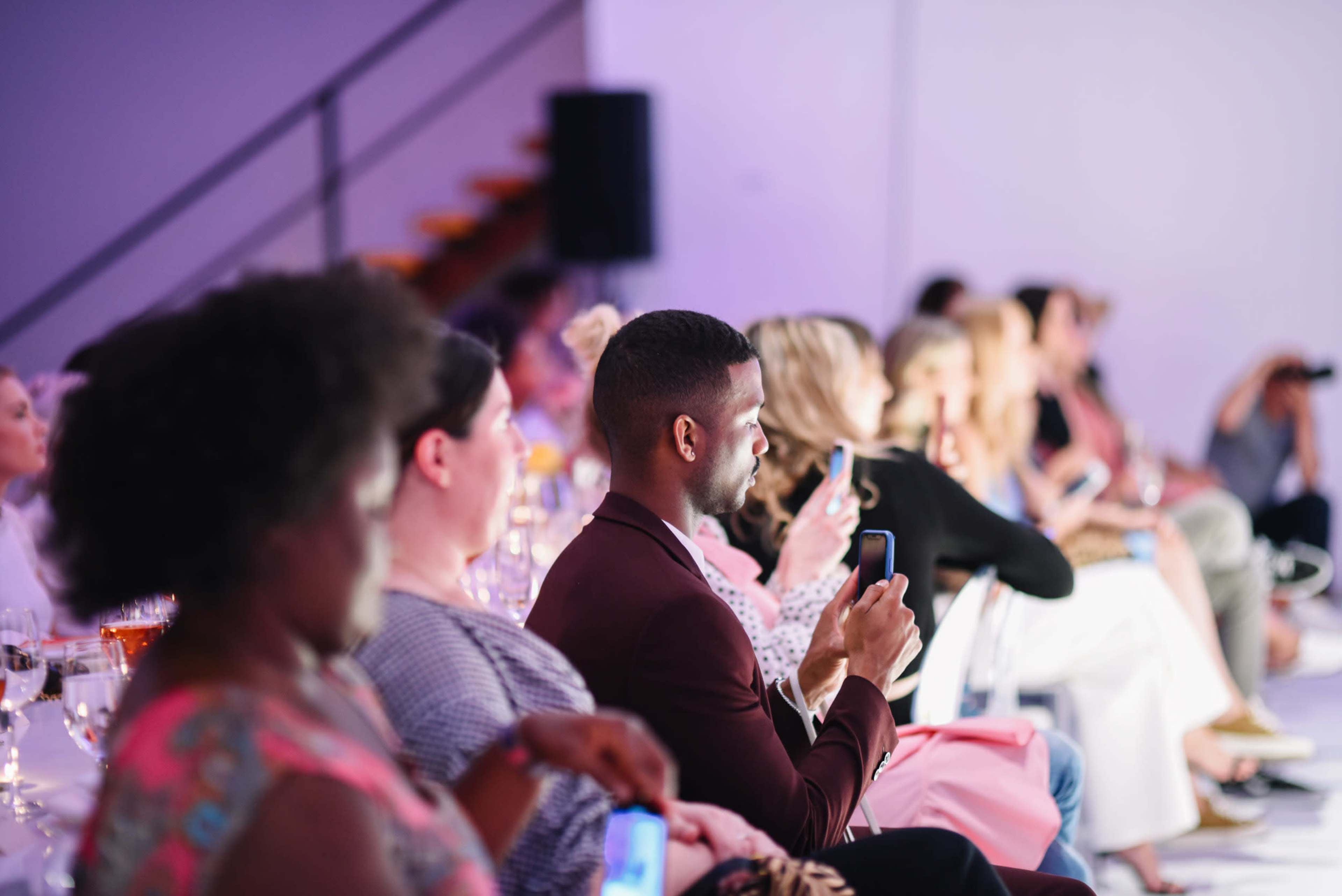 A diverse audience watches an event while some attendees use their phones to capture the moment.