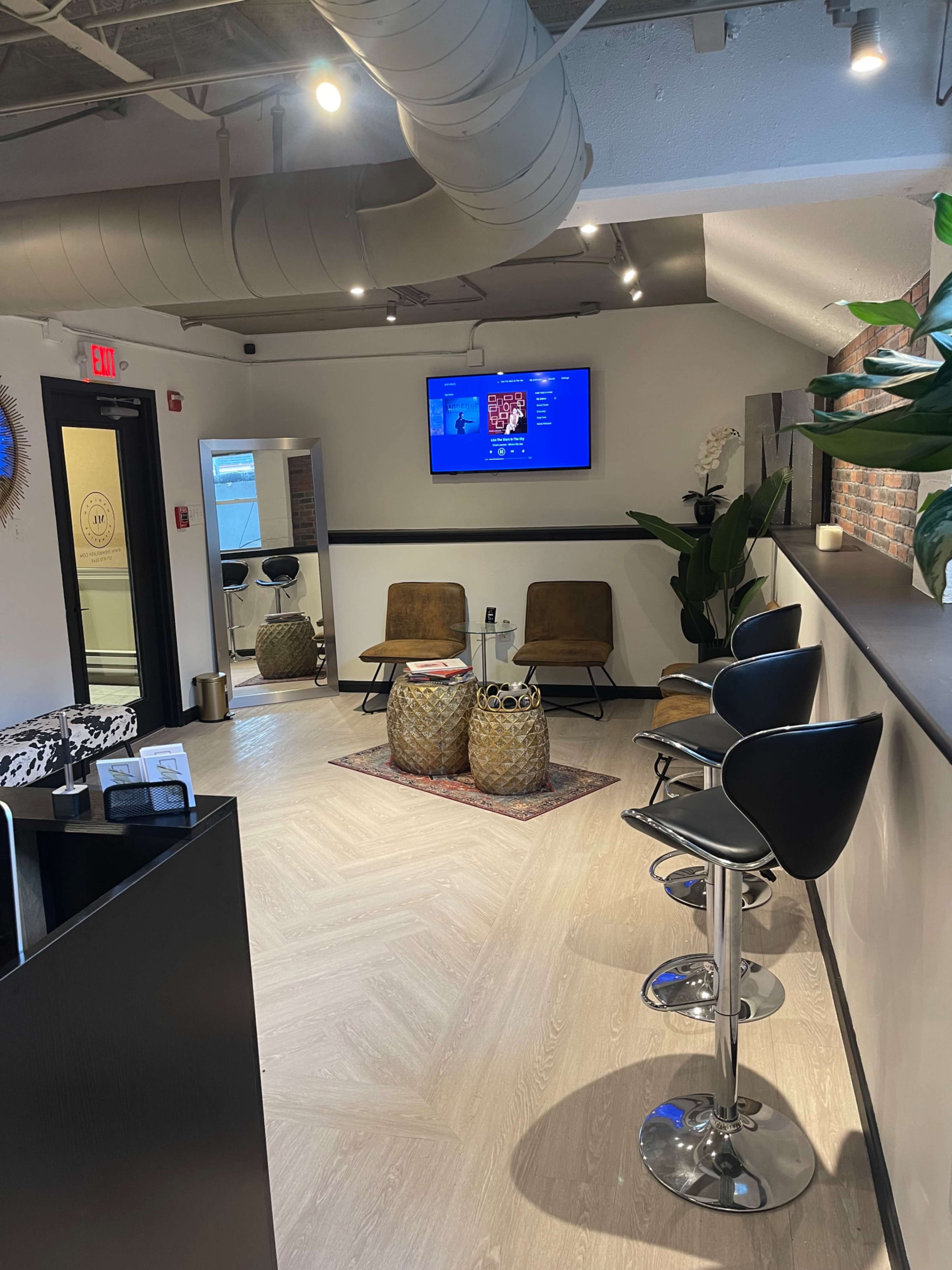 The image shows a modern waiting area with two chairs, a bar-style seating area, and decorative plants, featuring a wall-mounted television displaying content.
