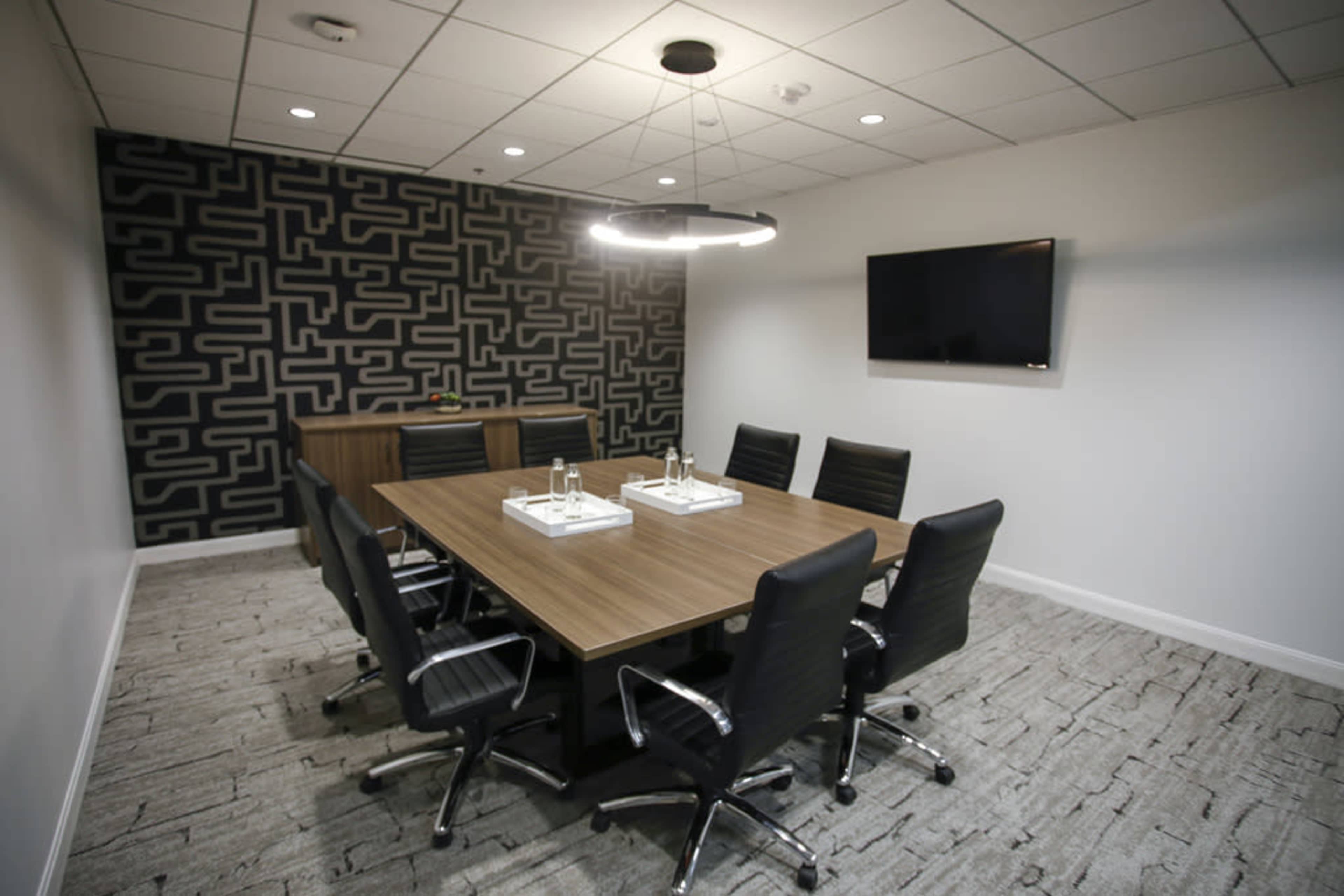A conference room features a long wooden table surrounded by black chairs, with a patterned wall and a television mounted on the opposite wall.