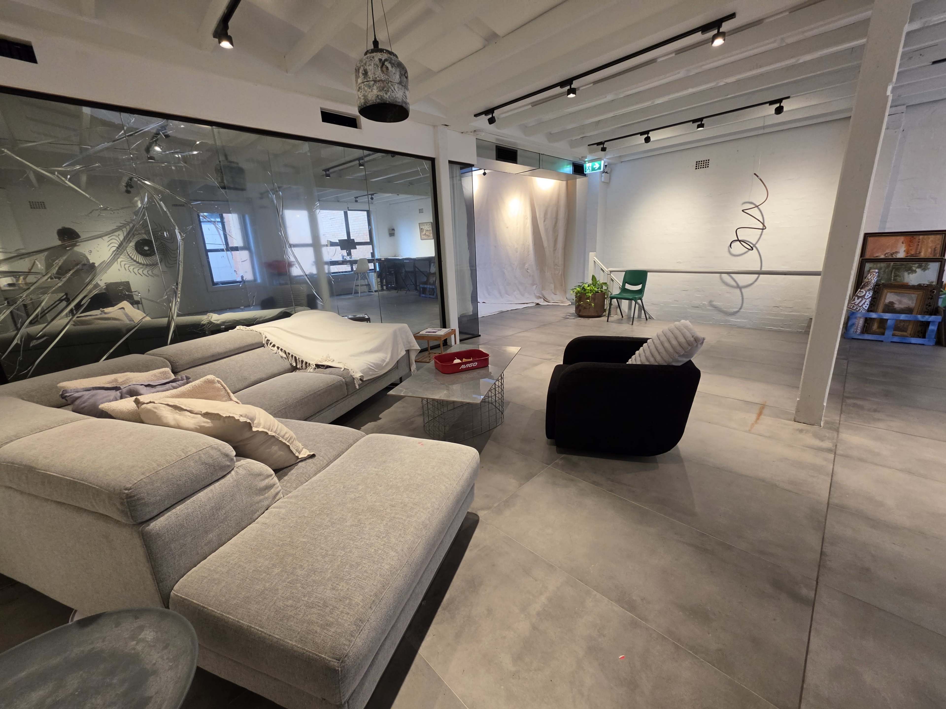 The image shows a modern, open space with a gray sofa, a black chair, and large windows, featuring a mix of contemporary art and plants.