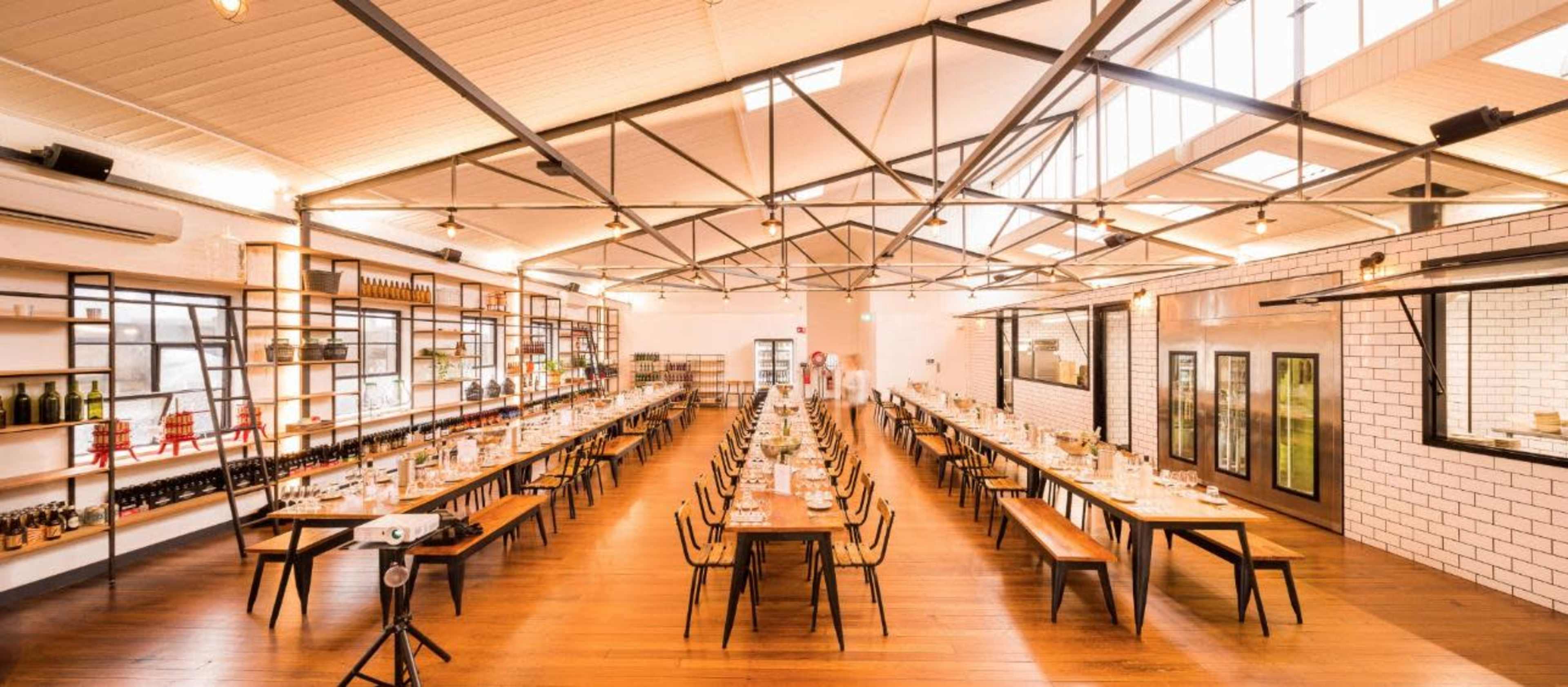A spacious dining area with long wooden tables set for a meal, illuminated by large windows and industrial-style lighting.