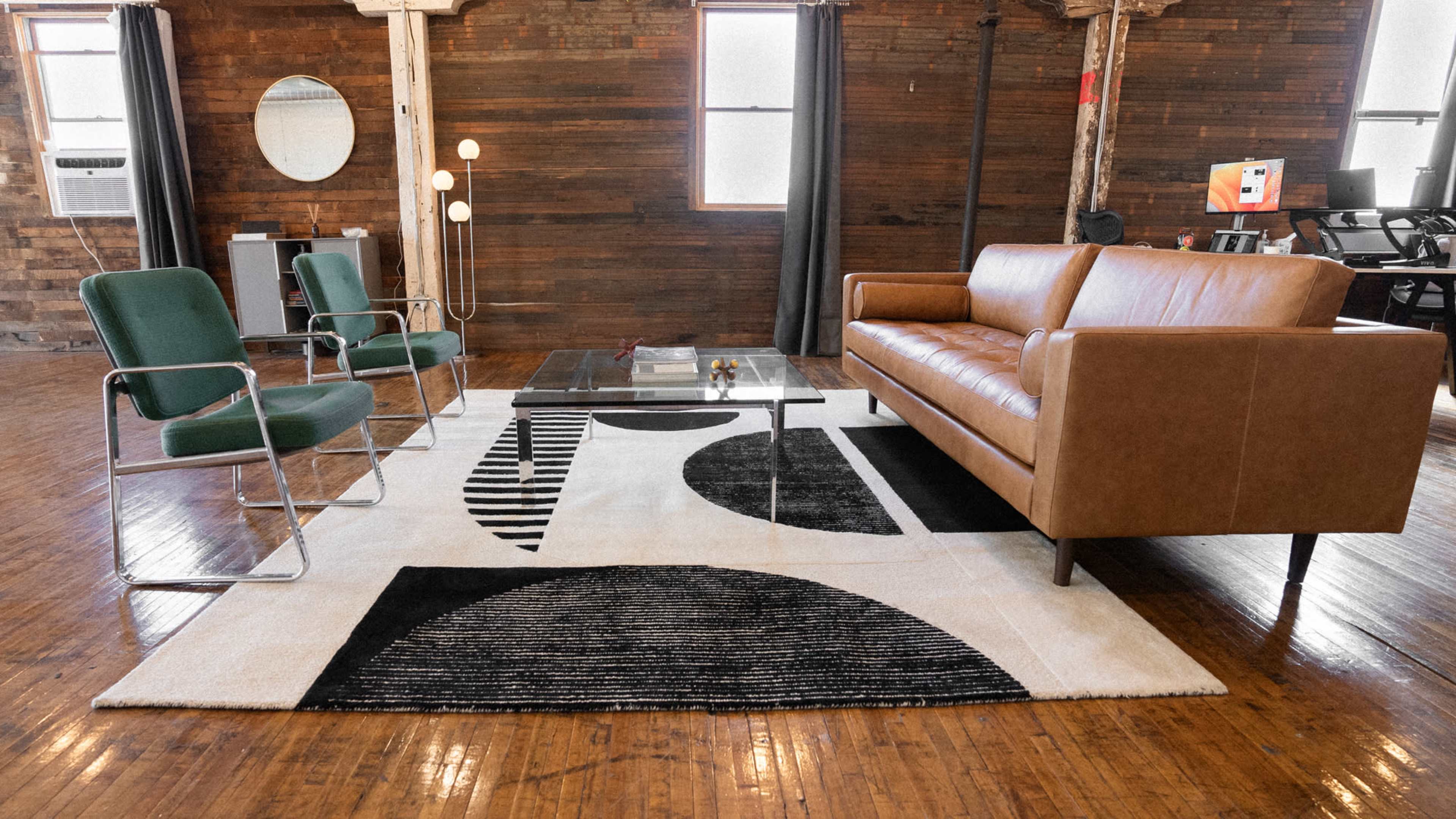 A living room with a brown leather sofa, two green chairs, a glass coffee table on a patterned rug, and wooden walls.