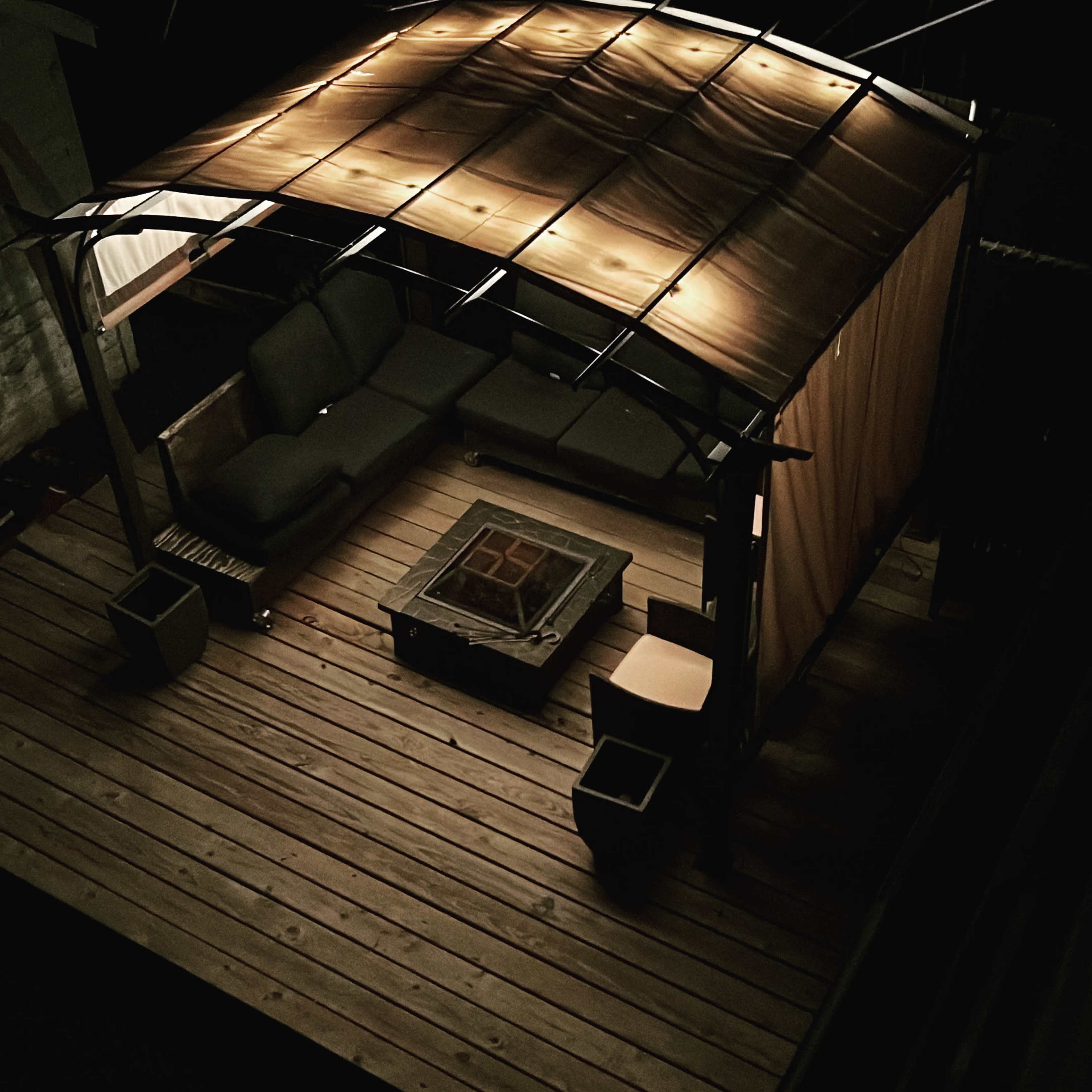 A wooden deck features a covered seating area with a sofa and a fire pit in the center, surrounded by potted plants.