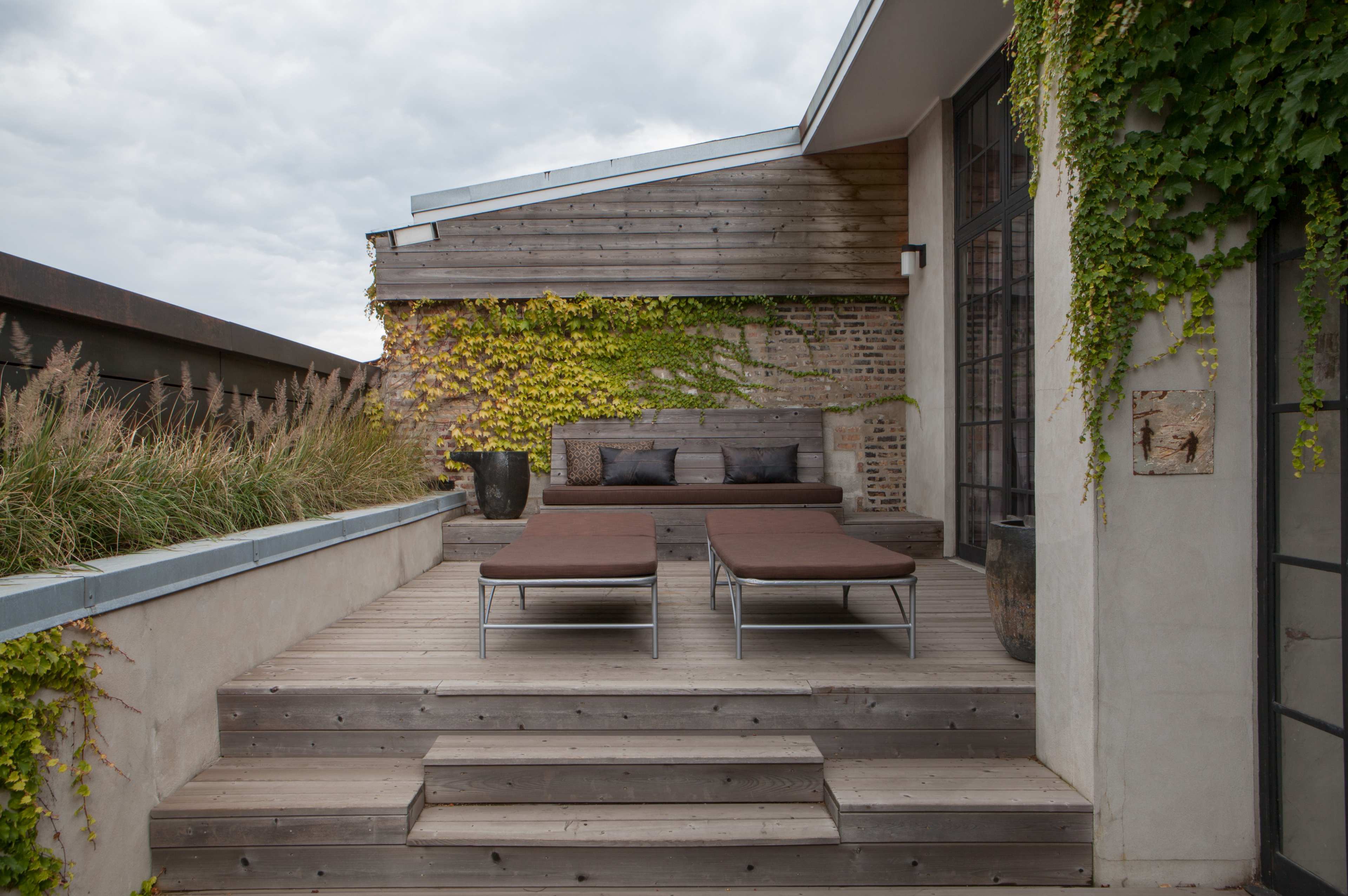 A terrace featuring wooden steps leading up to two brown lounge chairs surrounded by greenery and a textured wall.