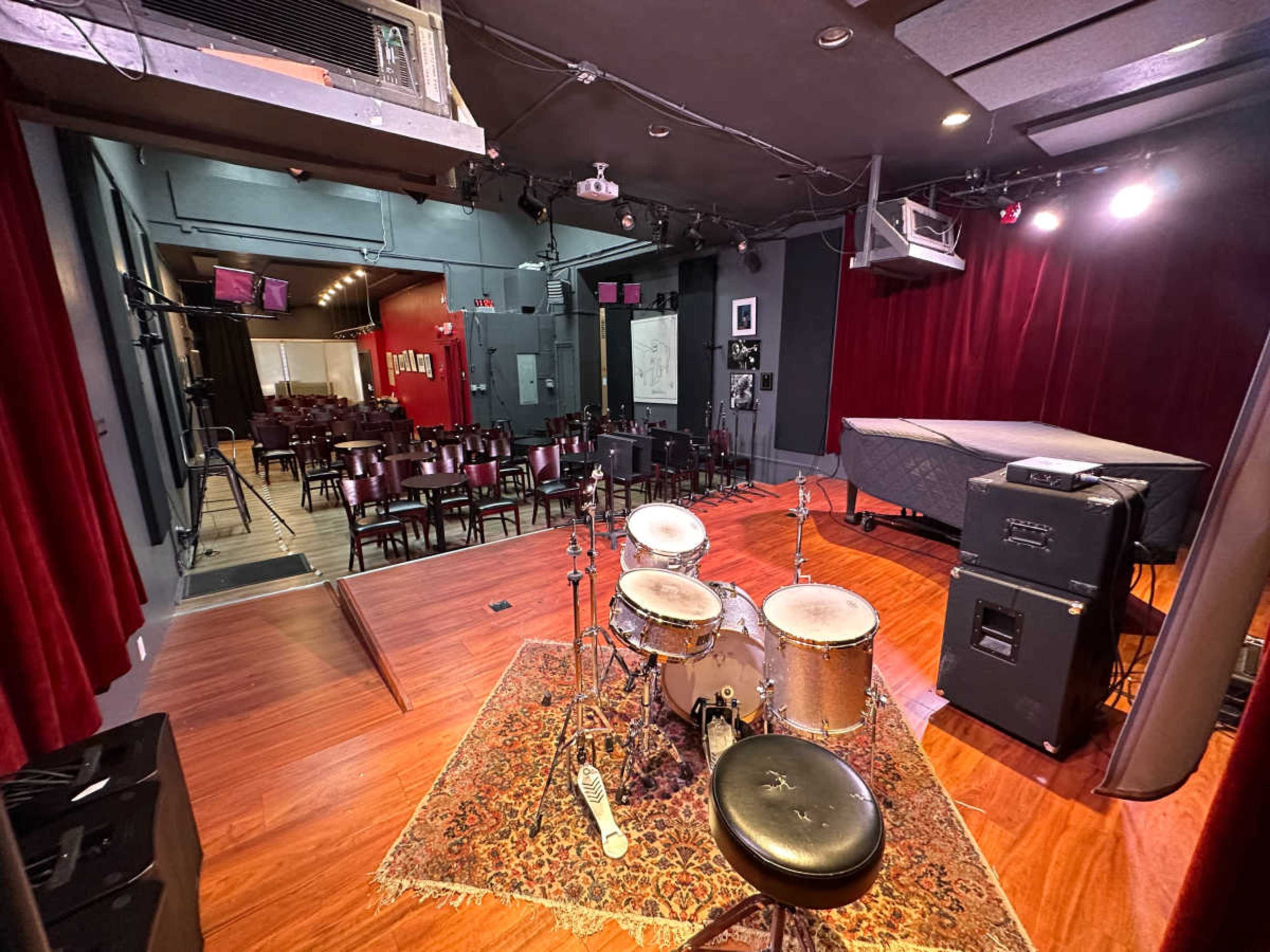 A performance space featuring a drum set on a wooden stage with a red velvet backdrop and a seating area visible in the background.