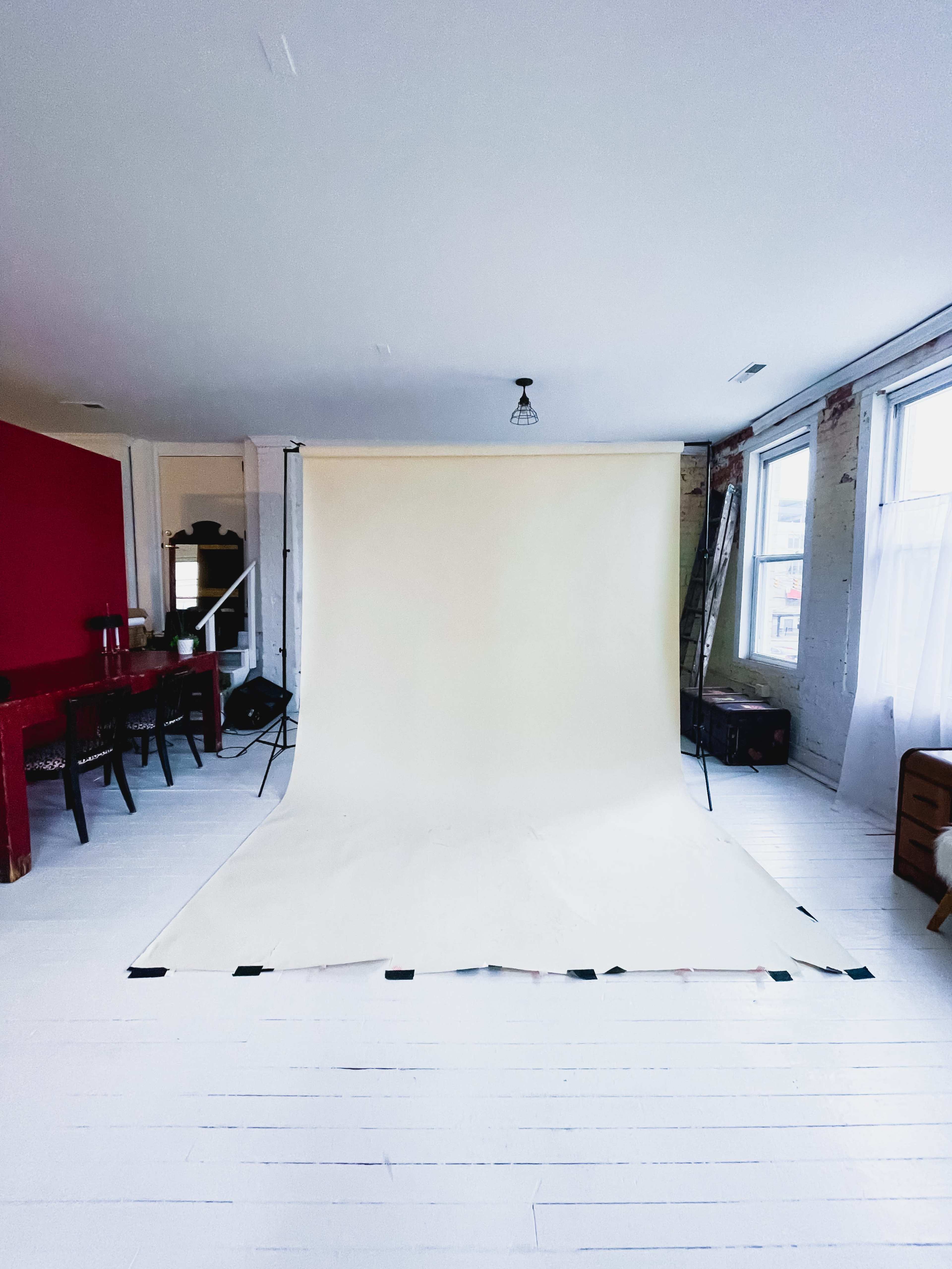 A photography studio featuring a large, neutral backdrop set on a white wooden floor, with a red wall visible in the background.