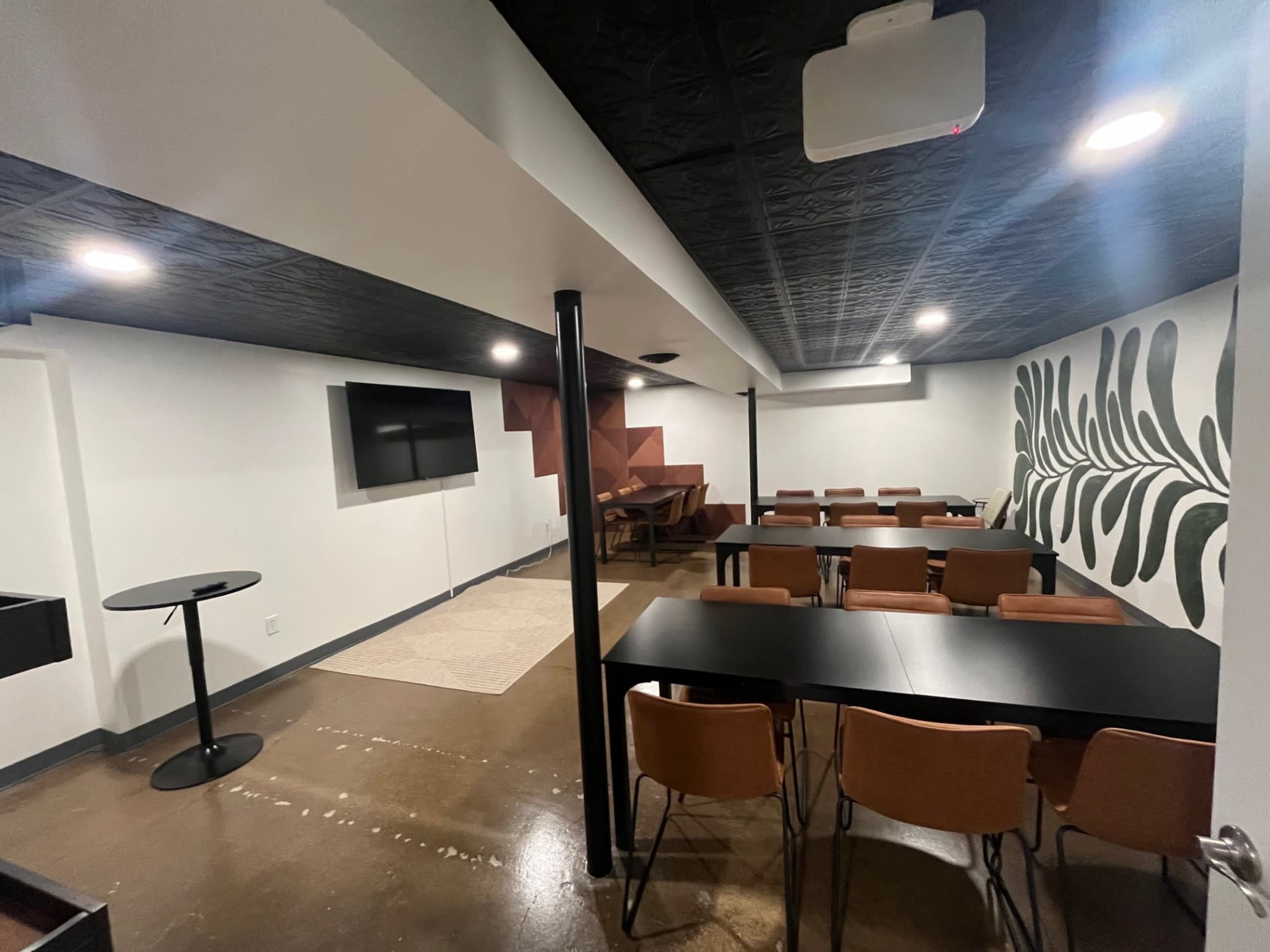 The image shows a modern meeting room equipped with a wall-mounted TV, several tables, chairs, and decorative wall elements.
