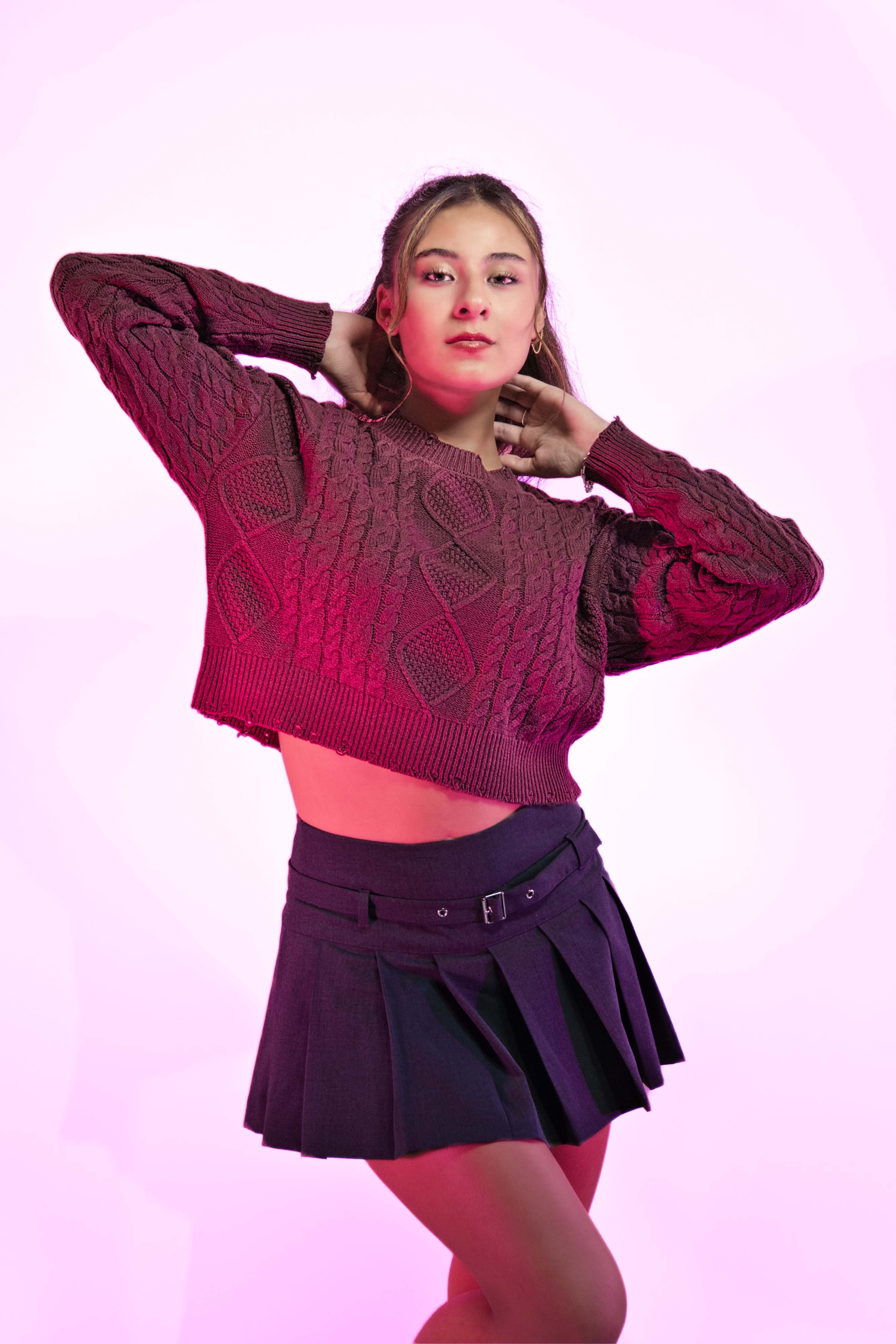 A young woman poses confidently in a cropped, textured sweater and a short pleated skirt against a pink background.