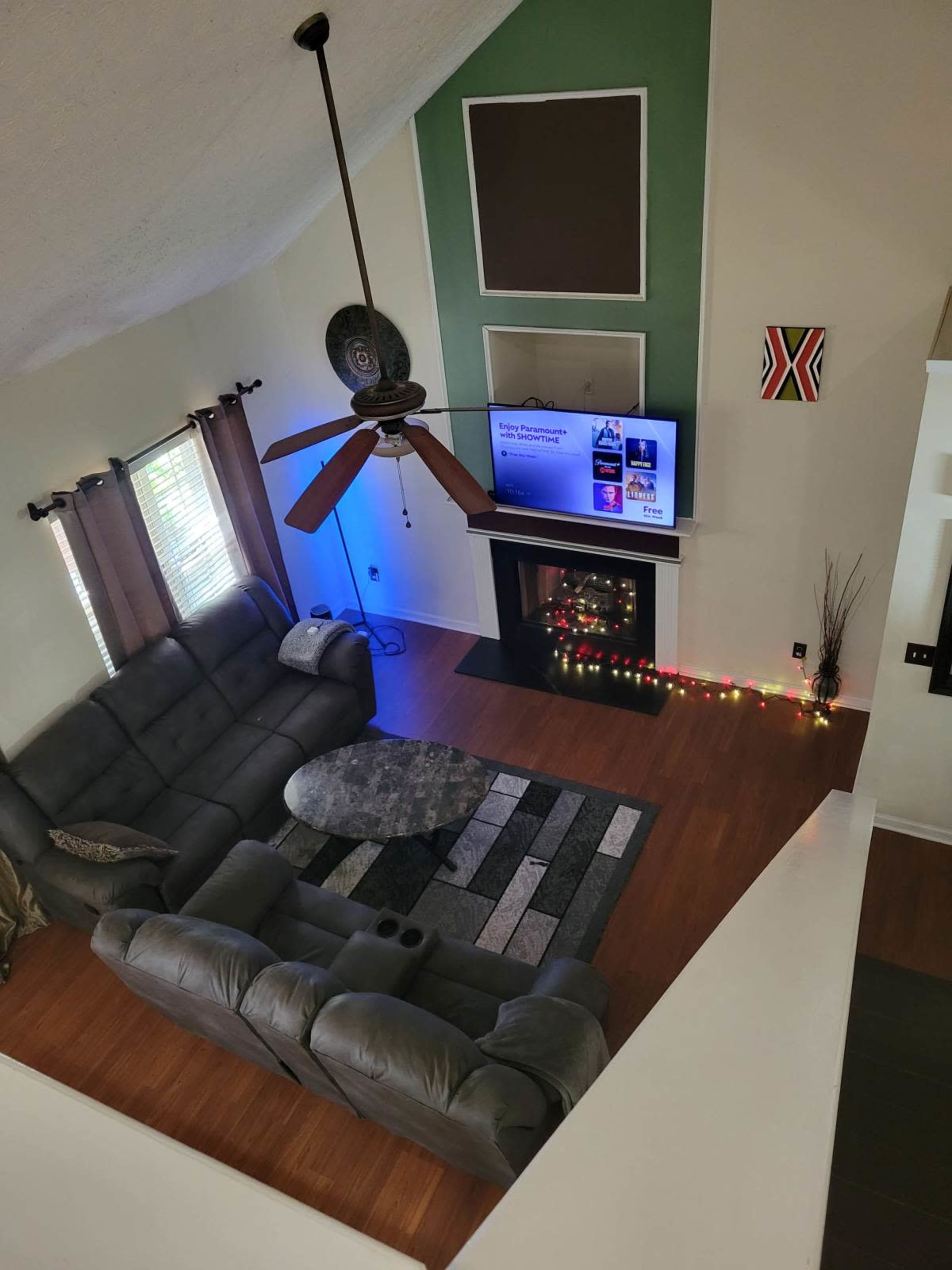 The image shows a spacious living room with a gray sectional sofa, a round coffee table on a rug, and a fireplace adorned with string lights, all under a ceiling fan.