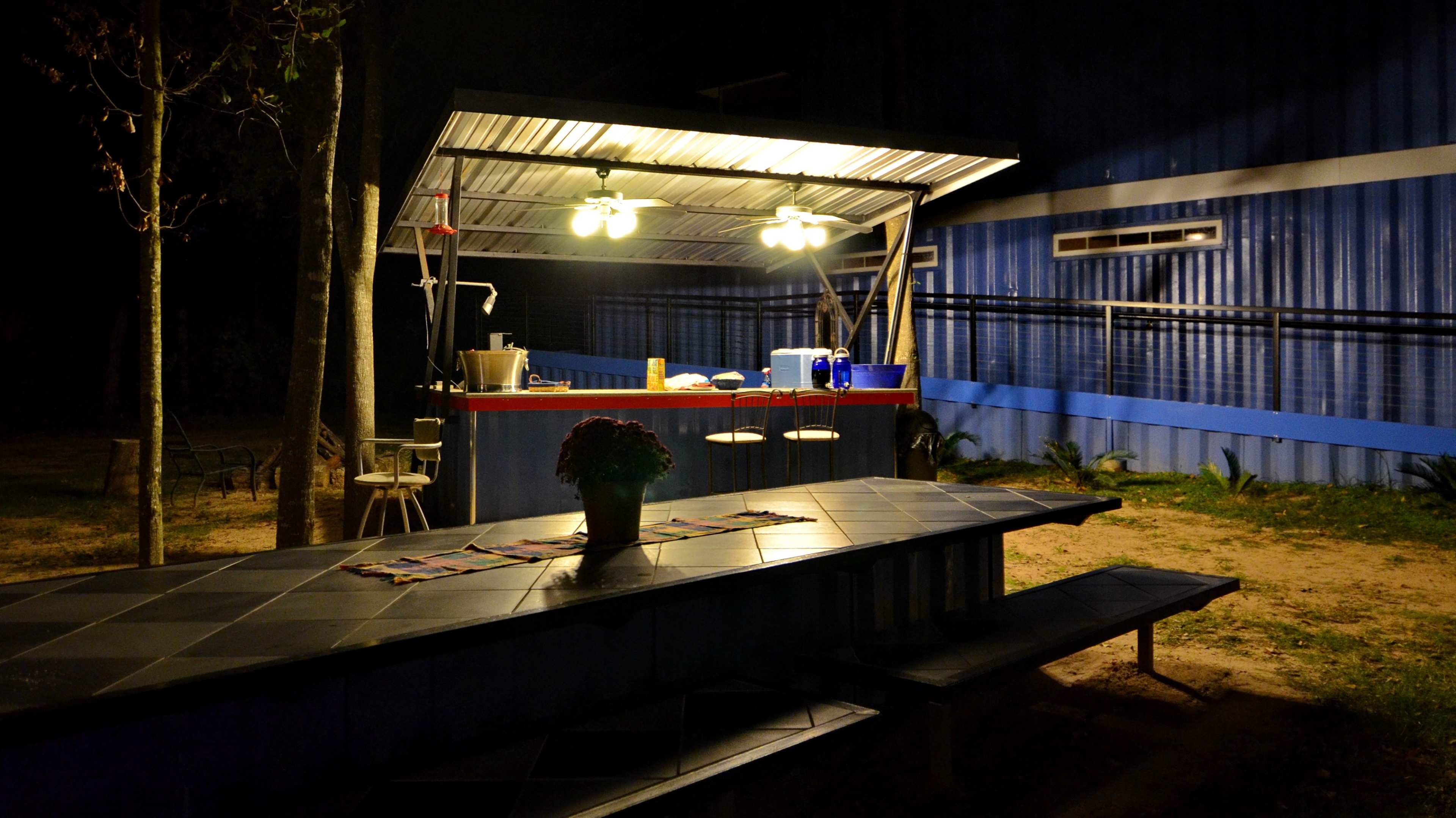 A brightly lit outdoor kitchen with a bar area and a nearby picnic table is set against a dark, wooded backdrop.