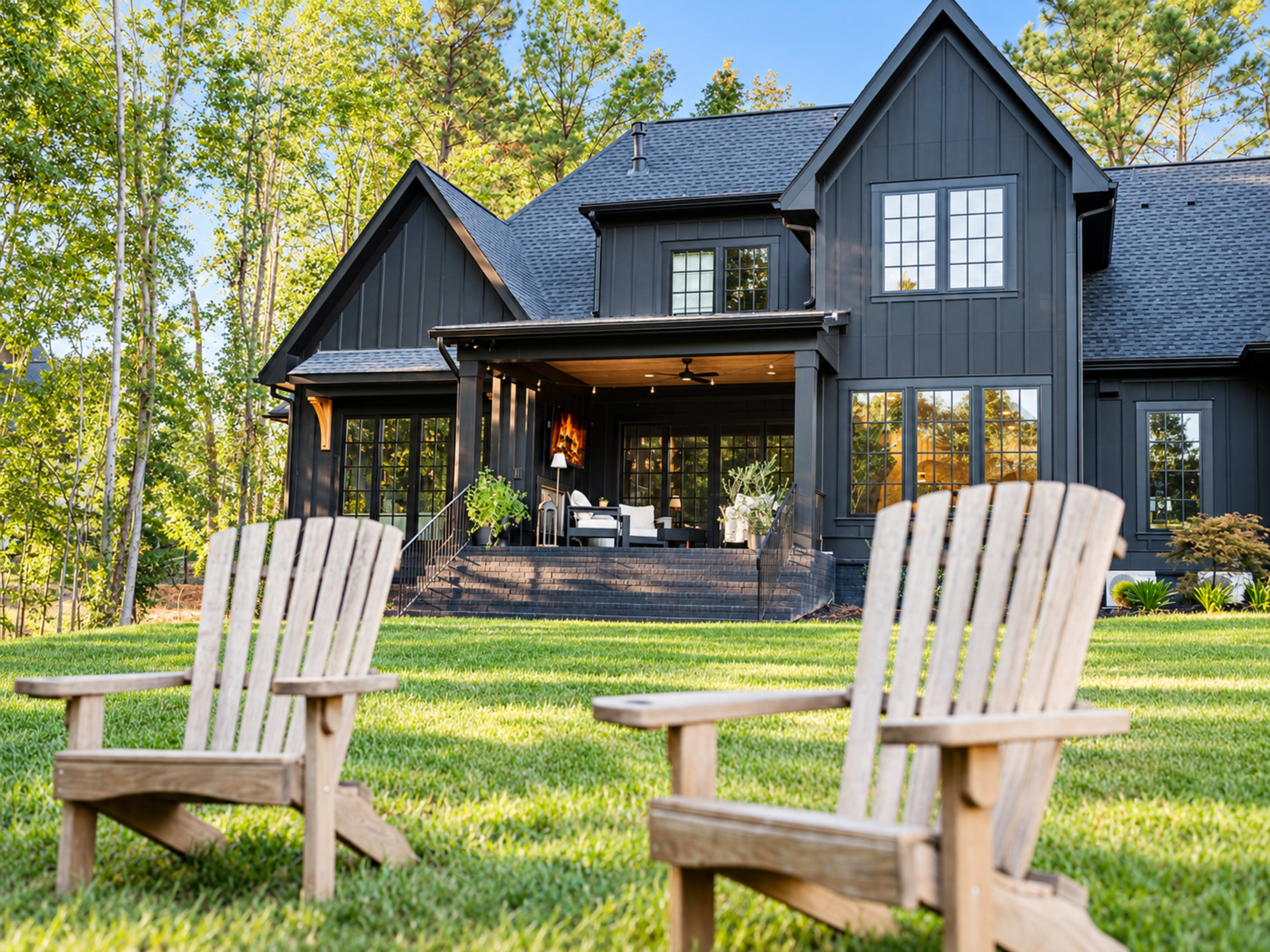 The image shows a modern black house with large windows and a spacious porch, surrounded by greenery, with two wooden Adirondack chairs in the foreground.