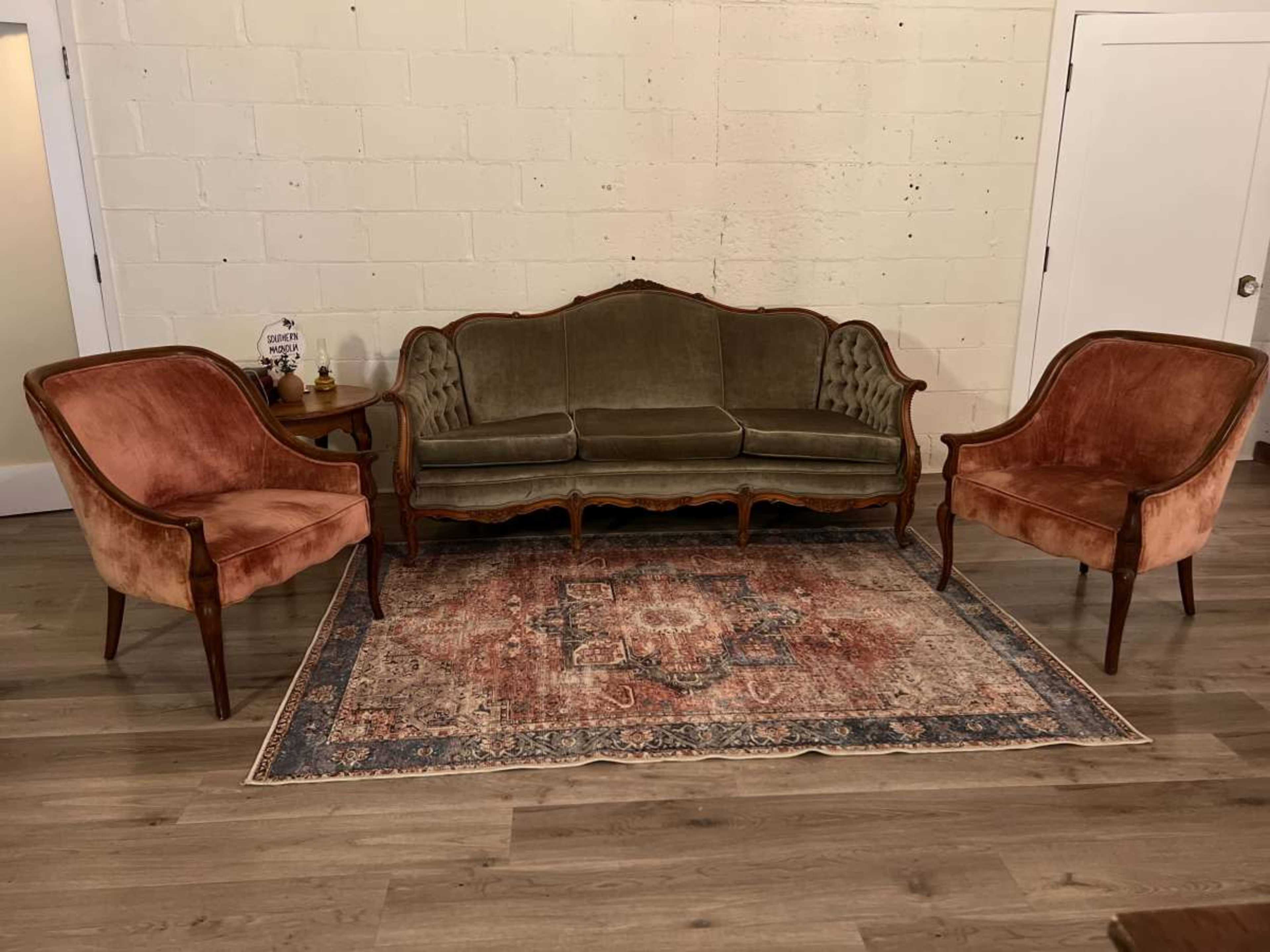 A vintage seating arrangement with a central green upholstered sofa flanked by two pink velvet chairs, all set on a patterned area rug against a plain wall.