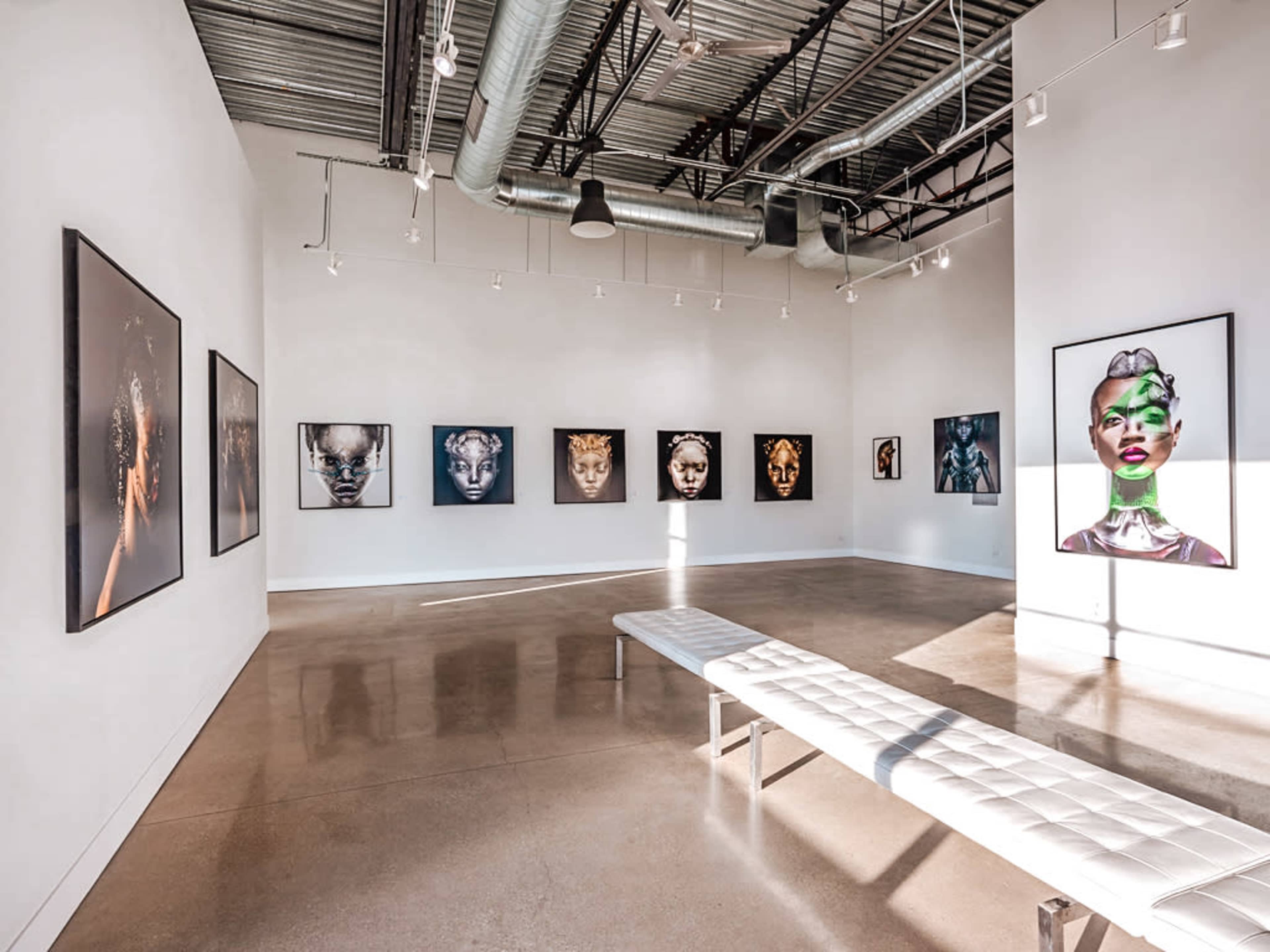The image depicts a modern art gallery featuring a series of framed portraits on the walls and a minimalist bench in the center.