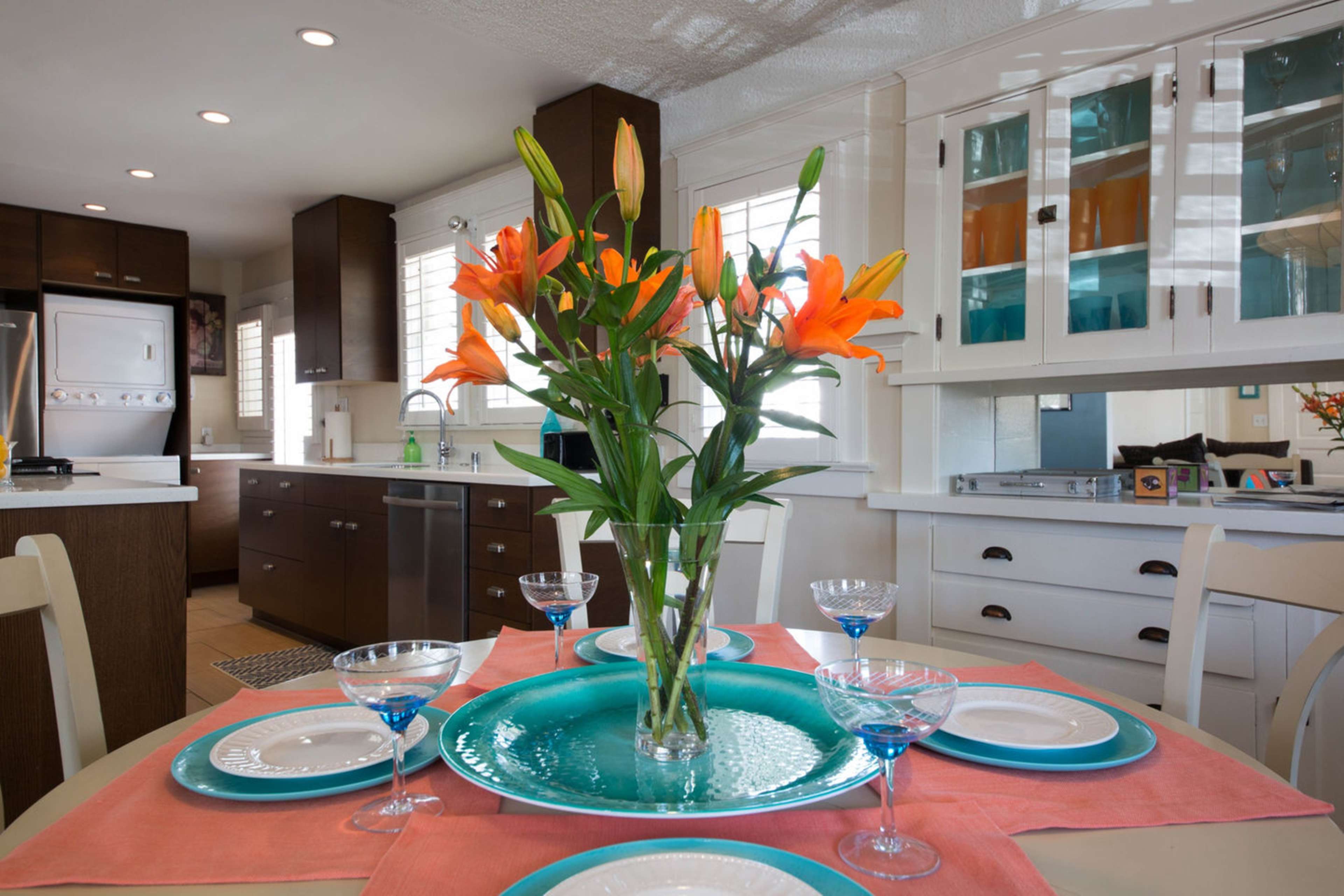 A dining table is set with plates and glasses, featuring a vase of orange lilies at the center, surrounded by a well-lit kitchen with dark cabinetry.