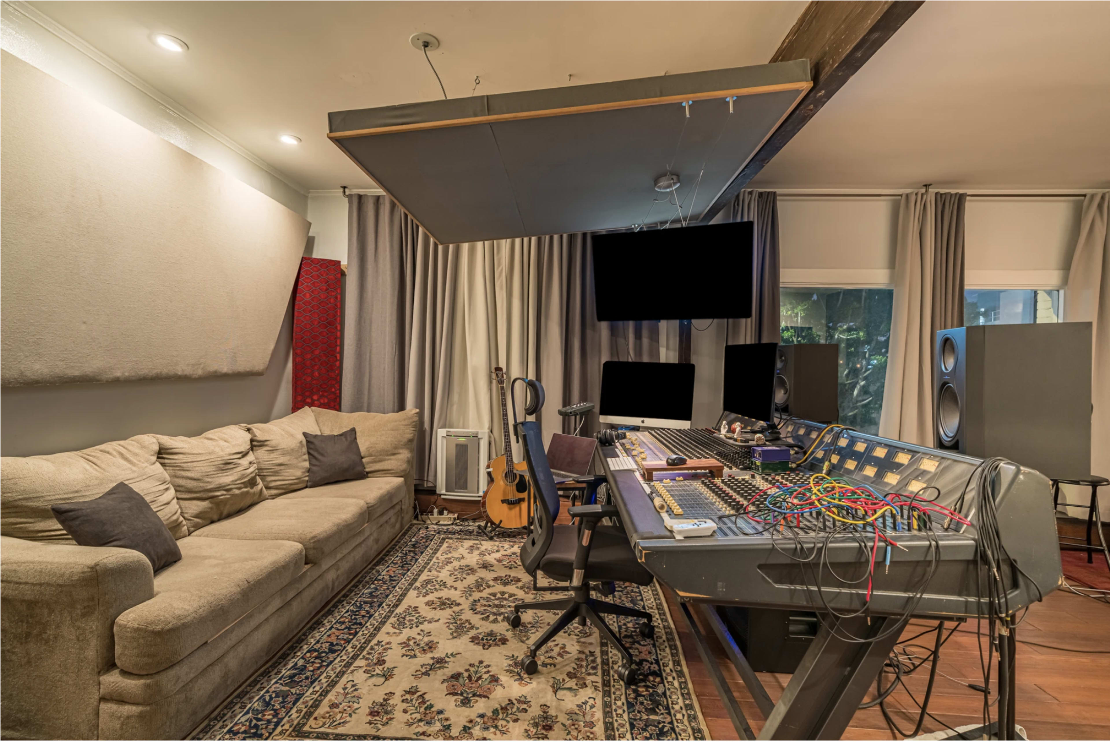 The image shows a music studio featuring a mixing console, a sofa, and musical instruments arranged in a room with acoustic panels and curtains.