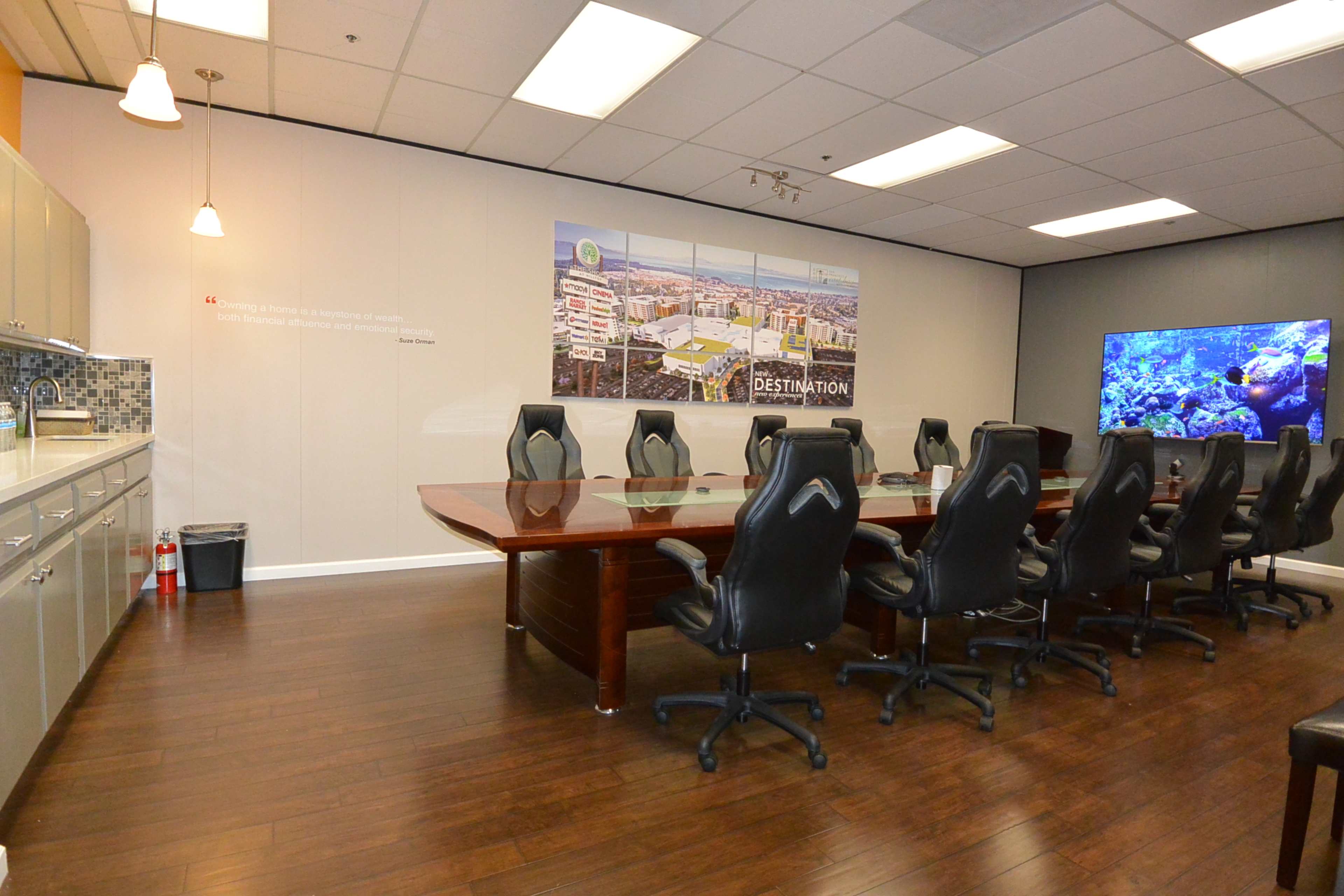 A conference room with a long wooden table surrounded by black executive chairs, a kitchenette area, and a large screen displaying underwater scenery.