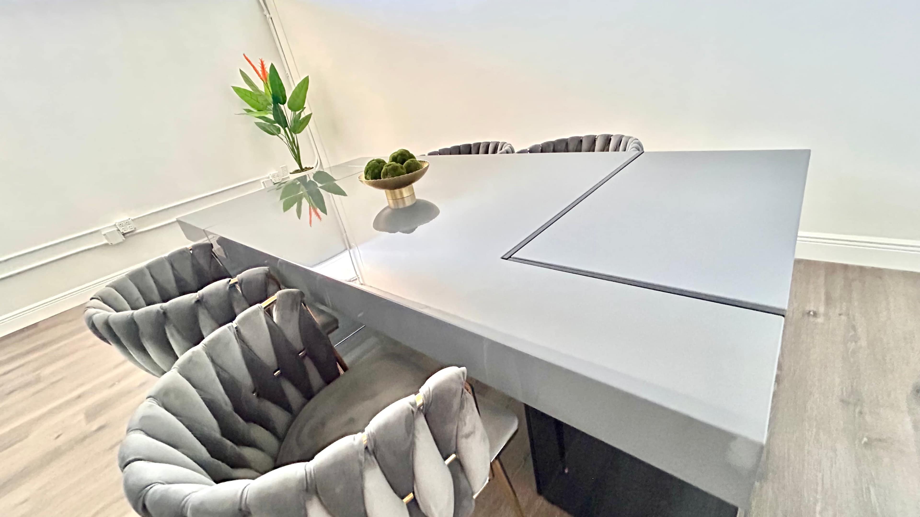 A modern dining table with a glossy surface is surrounded by tufted chairs, and a small vase with flowers and a bowl of decorative items sits atop the table.