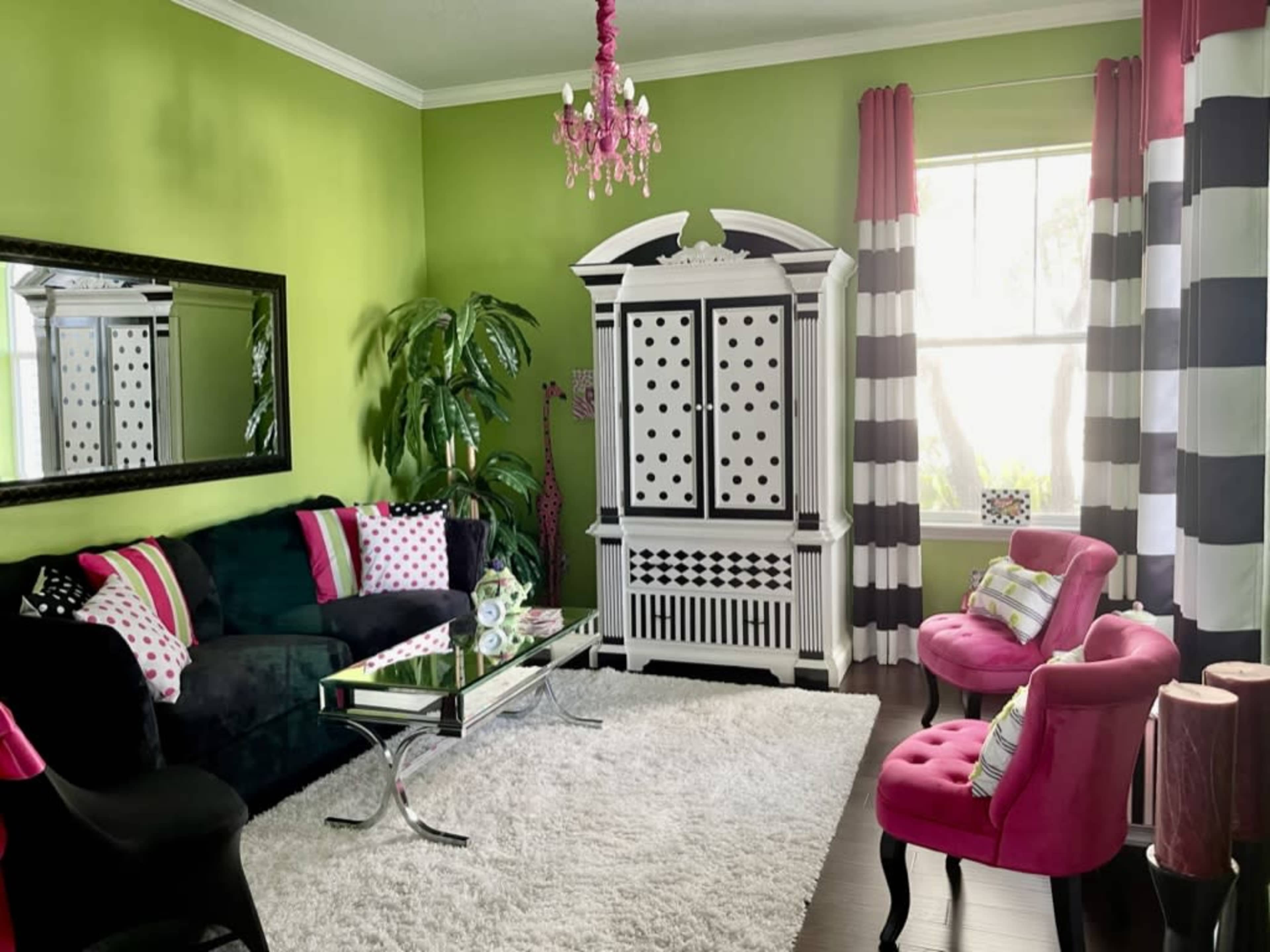 The room features green walls, a black velvet sofa with colorful pillows, a white cabinet with polka dots, and bright pink chairs, all set on a large white rug.