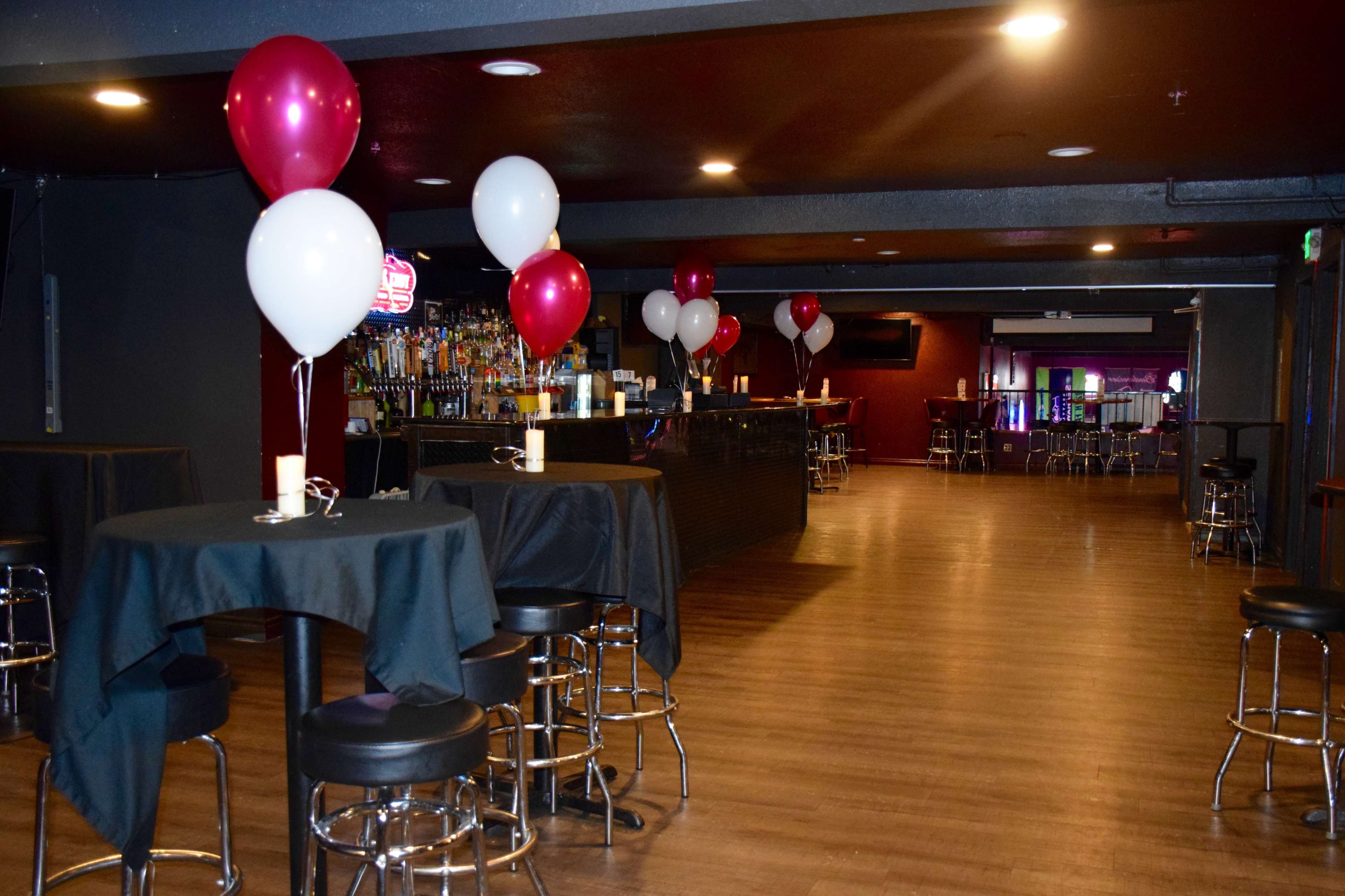 A dimly lit bar area with high-top tables covered in black cloths, featuring red and white balloons and a long bar lined with bottles in the background.