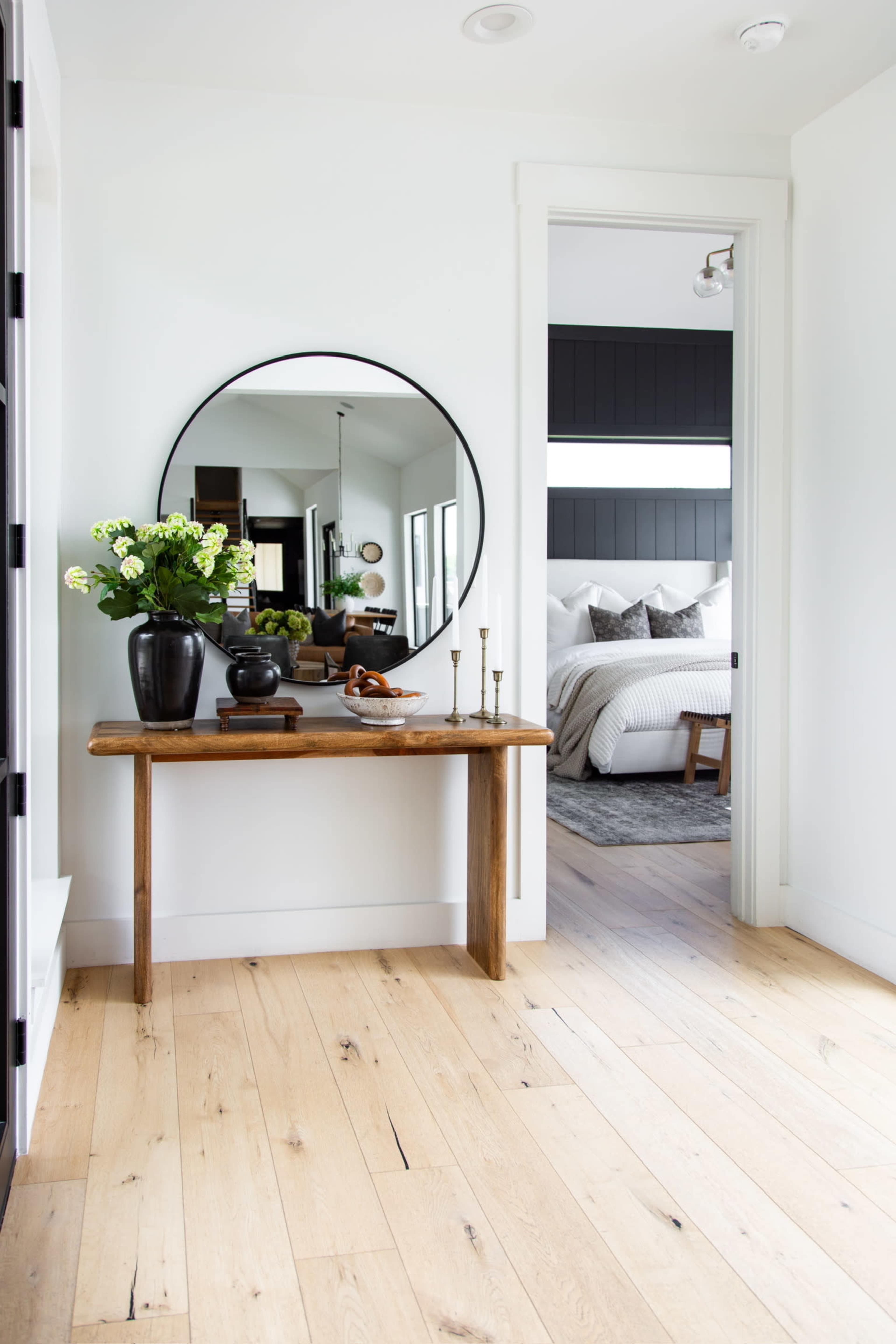 A bright entryway with a wooden console table, a round mirror, and a vase of flowers, leading into a bedroom area.