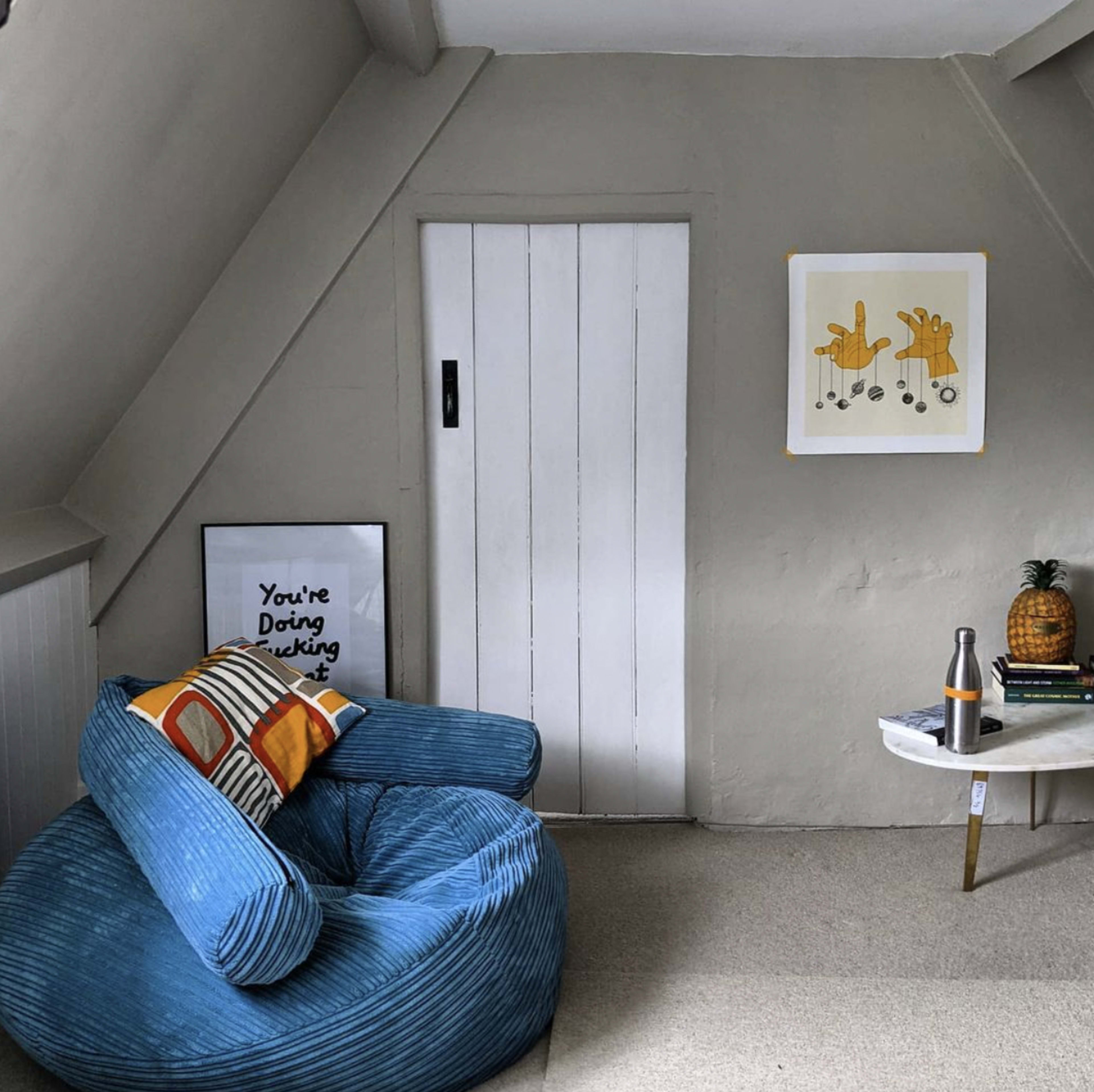A small, cozy room featuring a blue bean bag chair, a white door, a framed poster on the wall, and a small table with a pineapple and books.