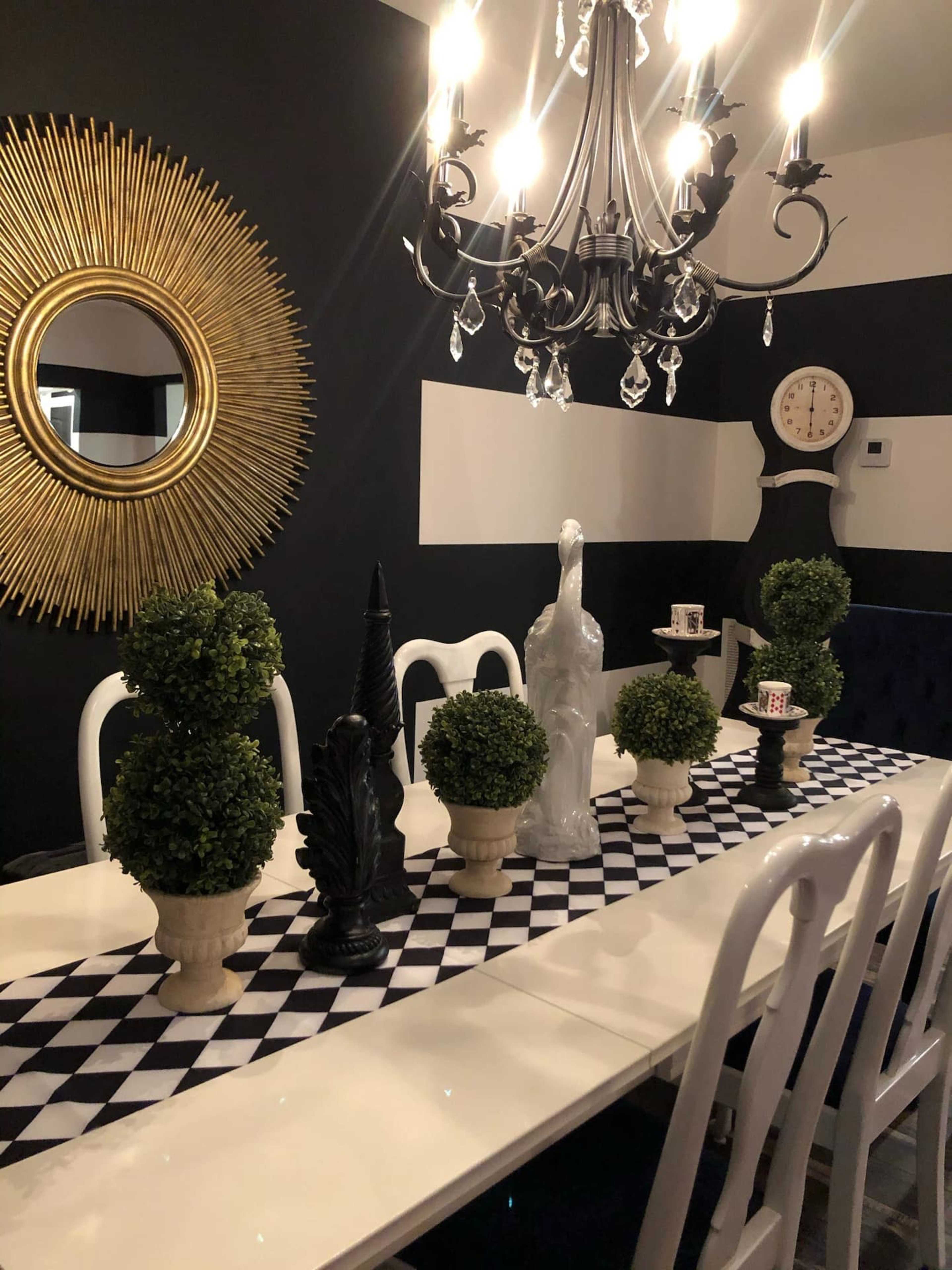 A dining table is set with a black and white checkered runner, surrounded by decorative topiaries and sculptures, under a chandelier and a sunburst mirror on the wall.