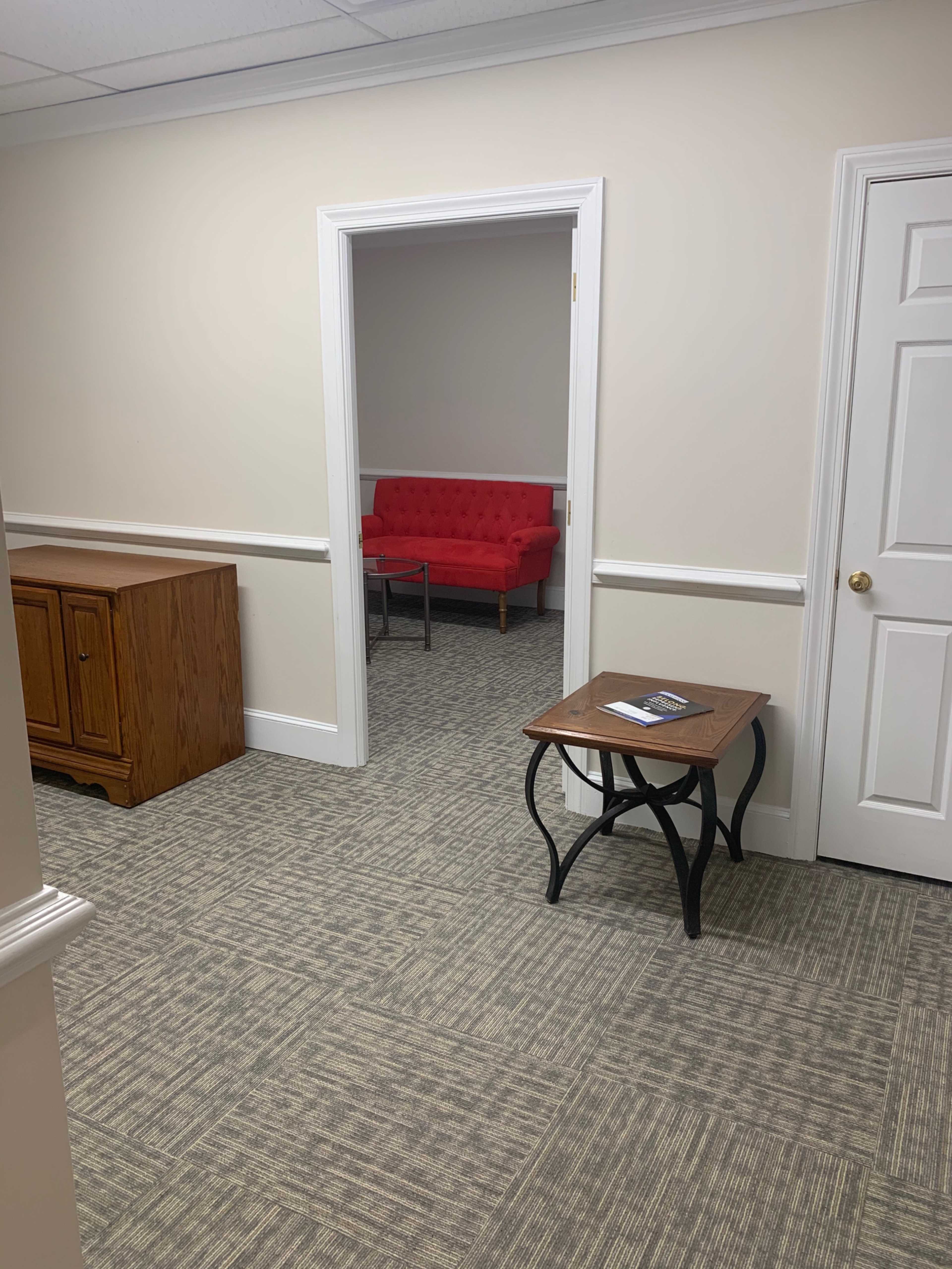 The image shows a hallway leading to a room with a red couch visible in the background, next to a wooden cabinet and a small table in the foreground.