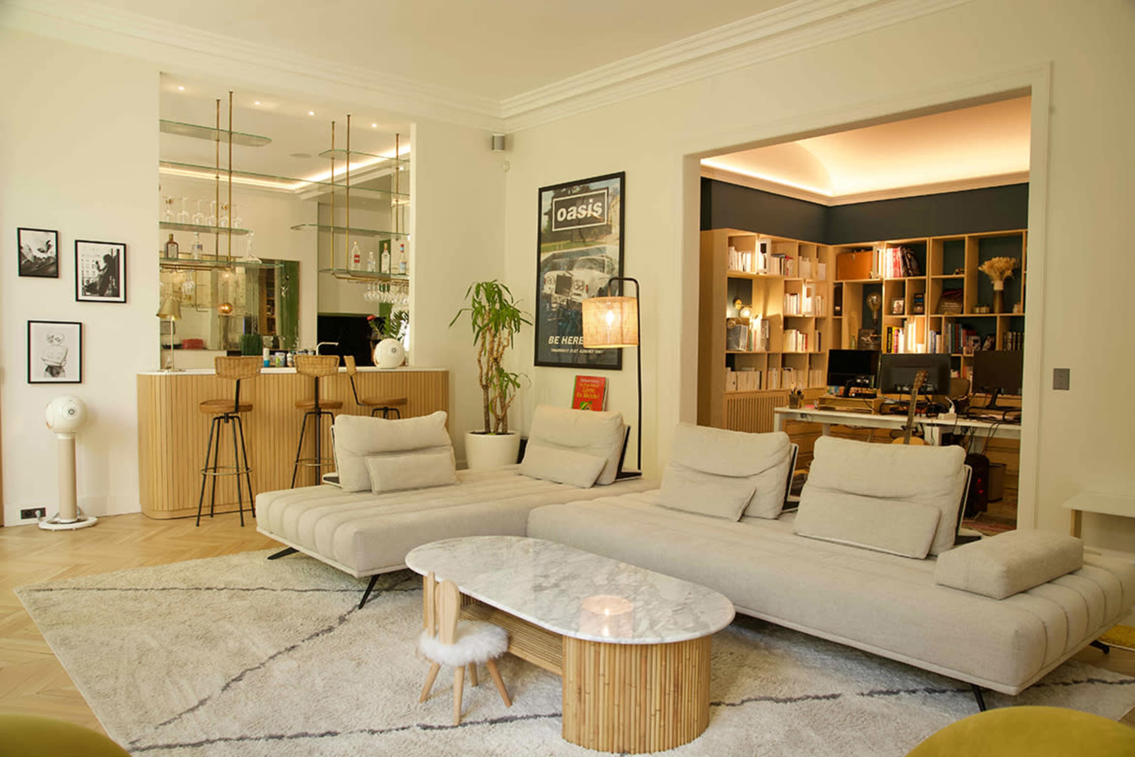 A modern living room features a pair of light-colored sofas, a round marble coffee table, a bamboo side table, and a view into a home office area with bookshelves.