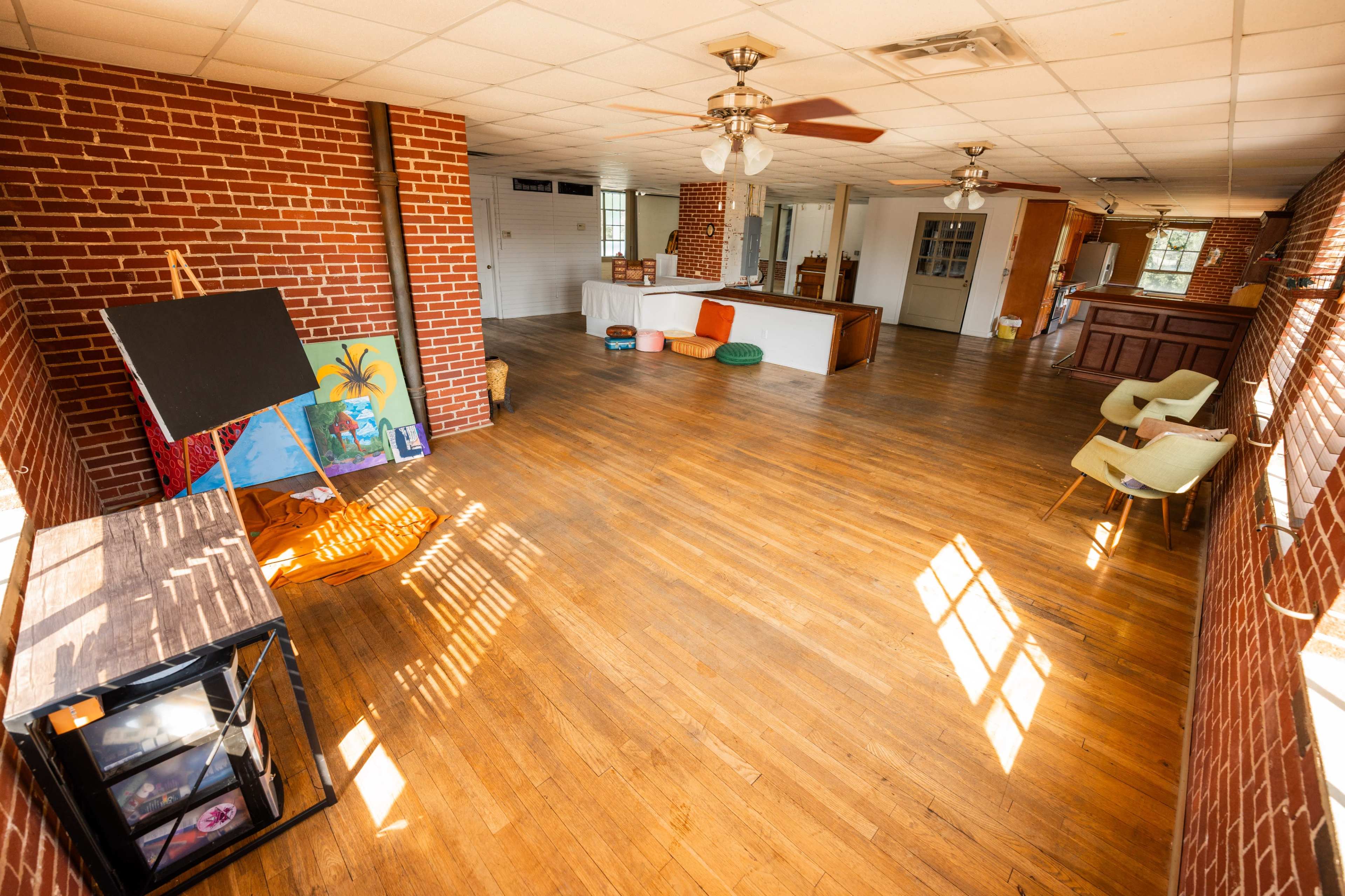 A spacious, empty room with wooden floors, brick walls, and large windows, featuring an easel with artwork in one corner and two chairs on the right.