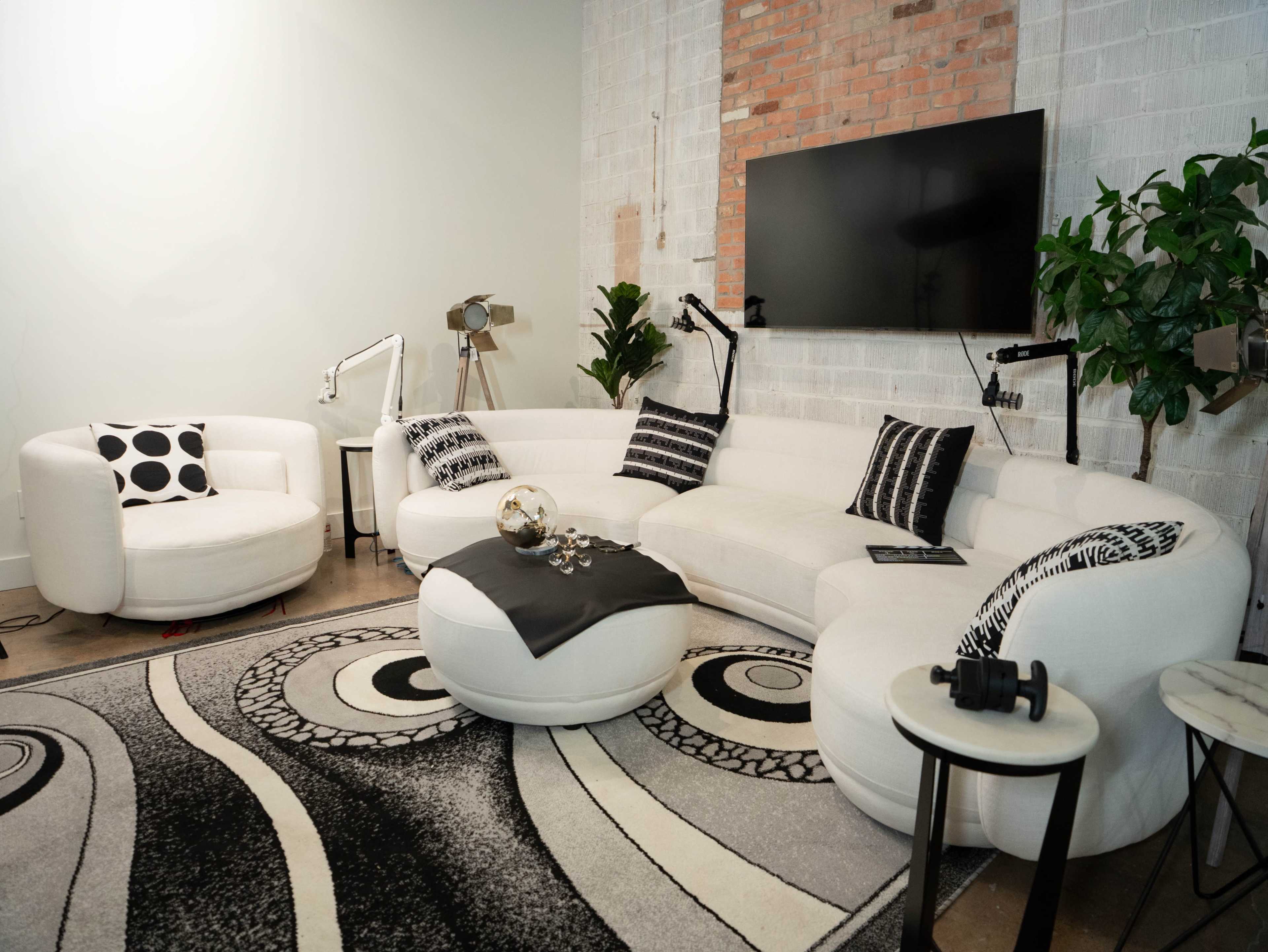 A modern living room features a curved white sectional sofa, a round accent chair, a large area rug with a bold black and white pattern, and a wall-mounted television, complemented by potted plants and stylish lighting.