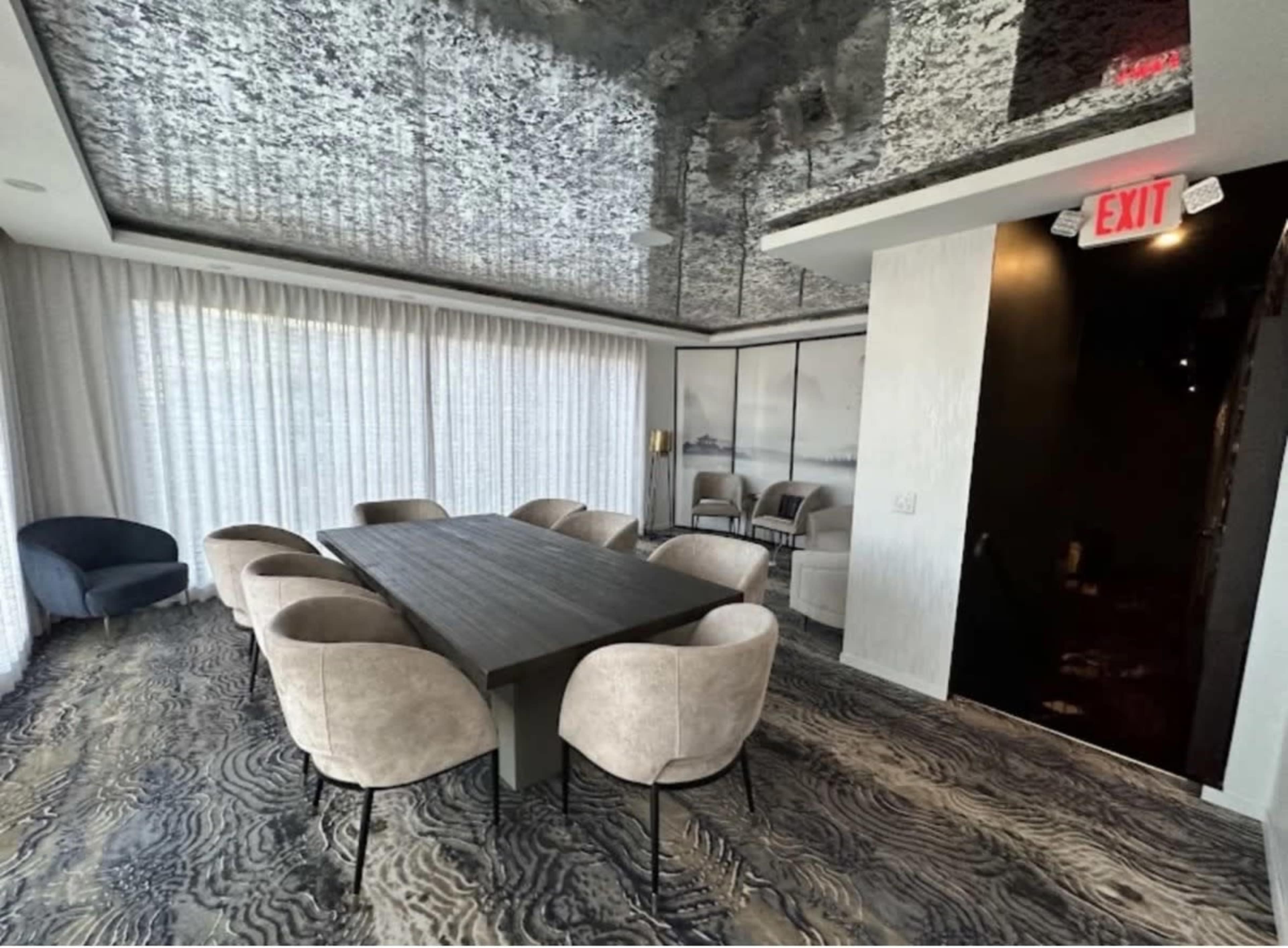 A modern conference room features a long table surrounded by upholstered chairs, with large windows covered by sheer drapes and a reflective ceiling.