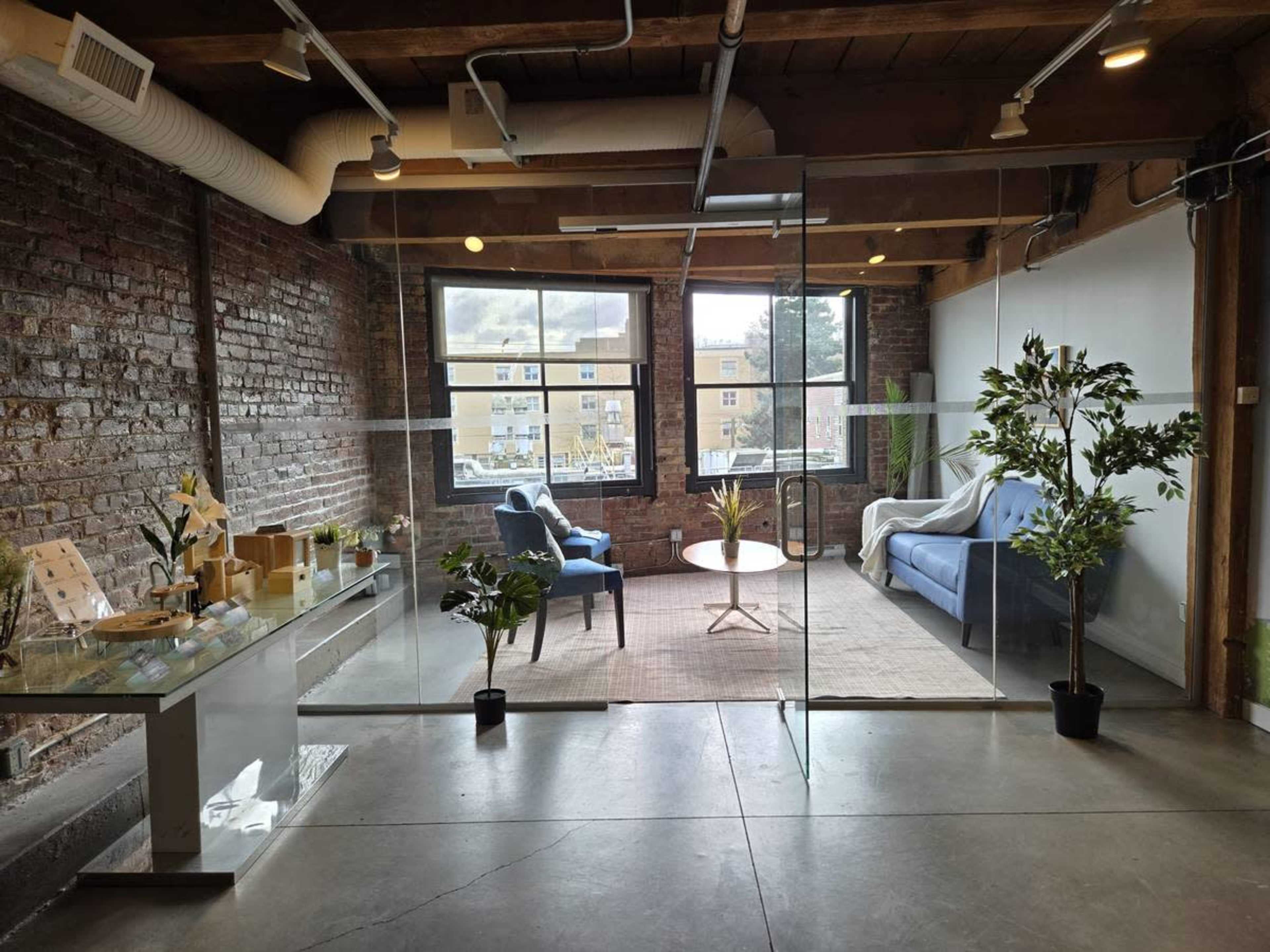 The image displays a modern office space featuring a glass partition with a seating area that includes two chairs, a coffee table, and decorative plants.
