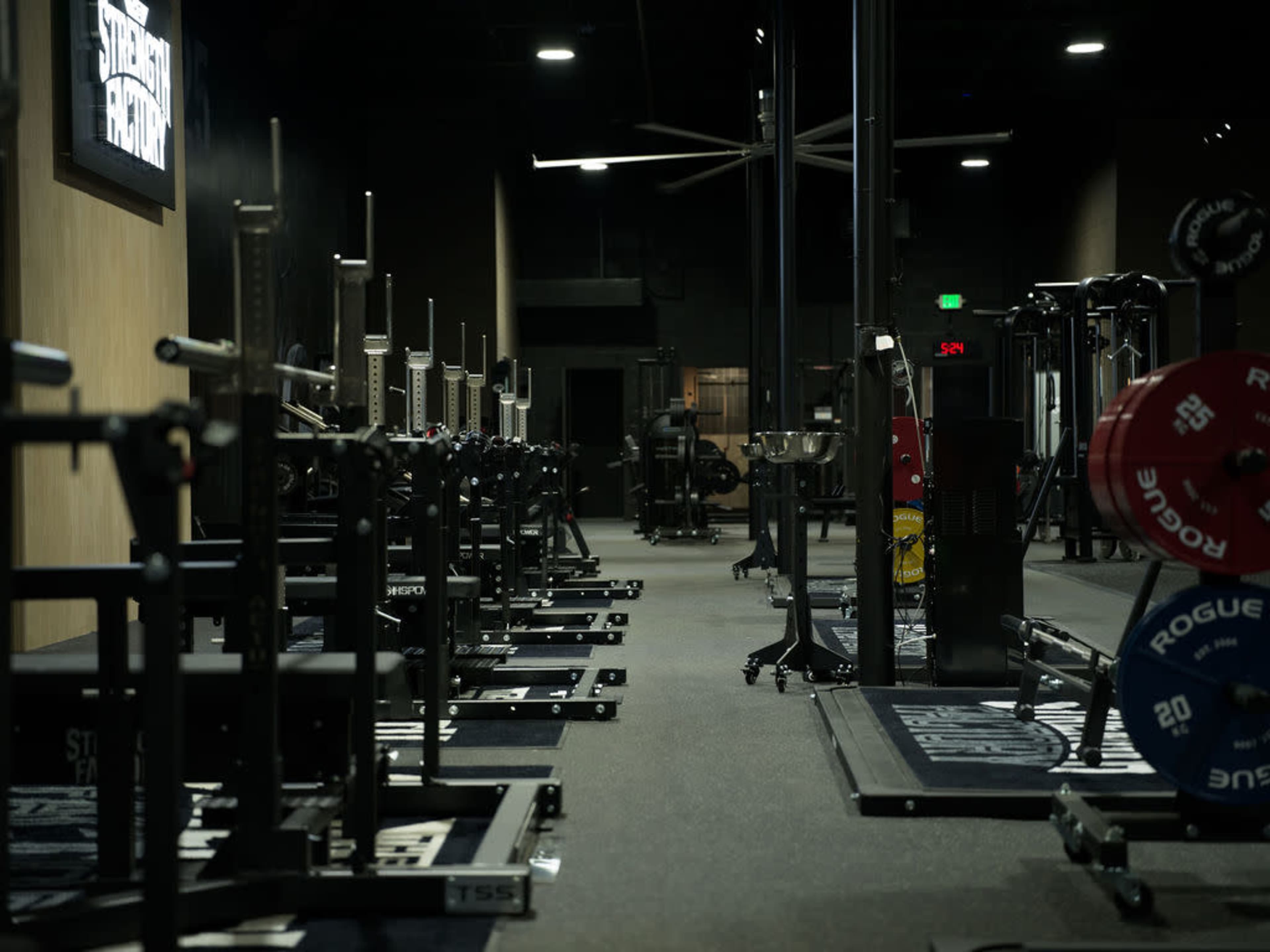 The image shows a dimly lit gym with various weightlifting equipment lined up along both sides of the space.