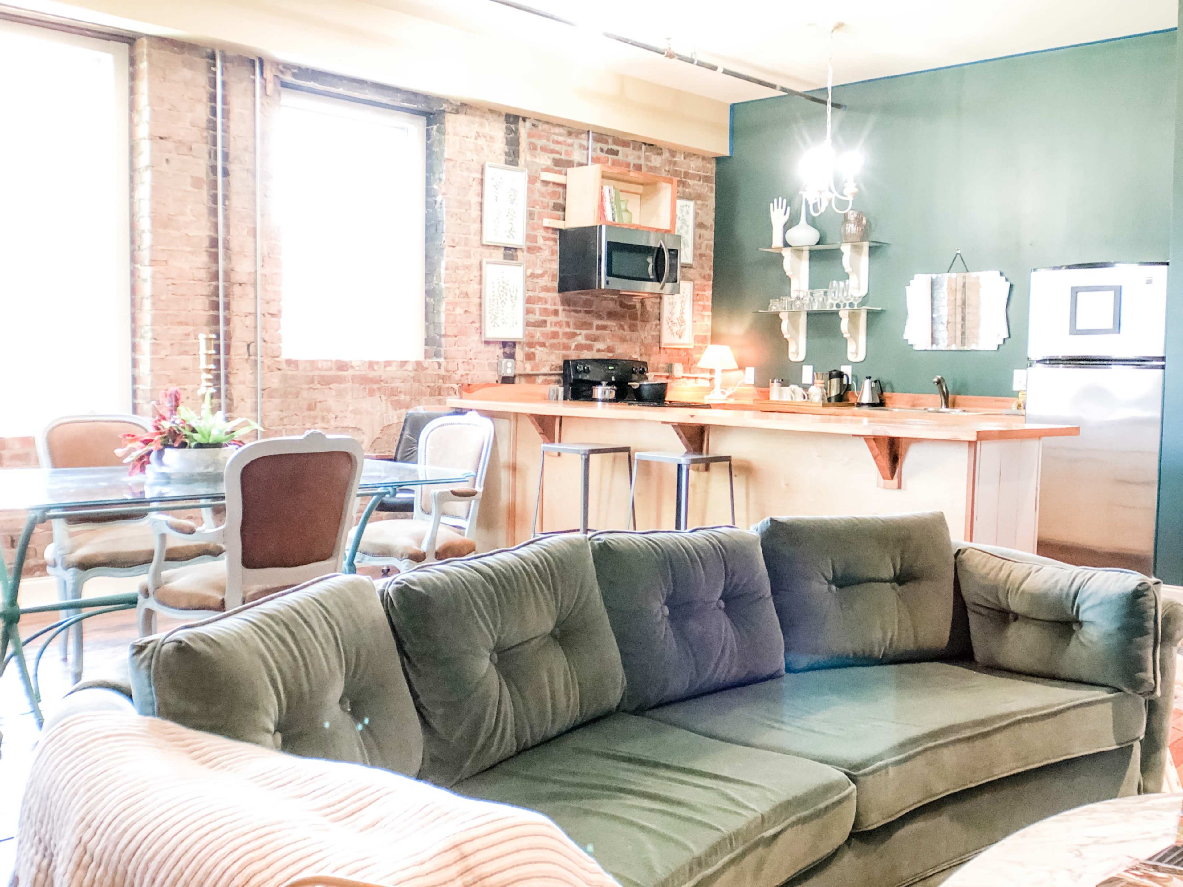 A cozy living area with a green velvet sofa, a dining table, and a kitchen counter against exposed brick walls.