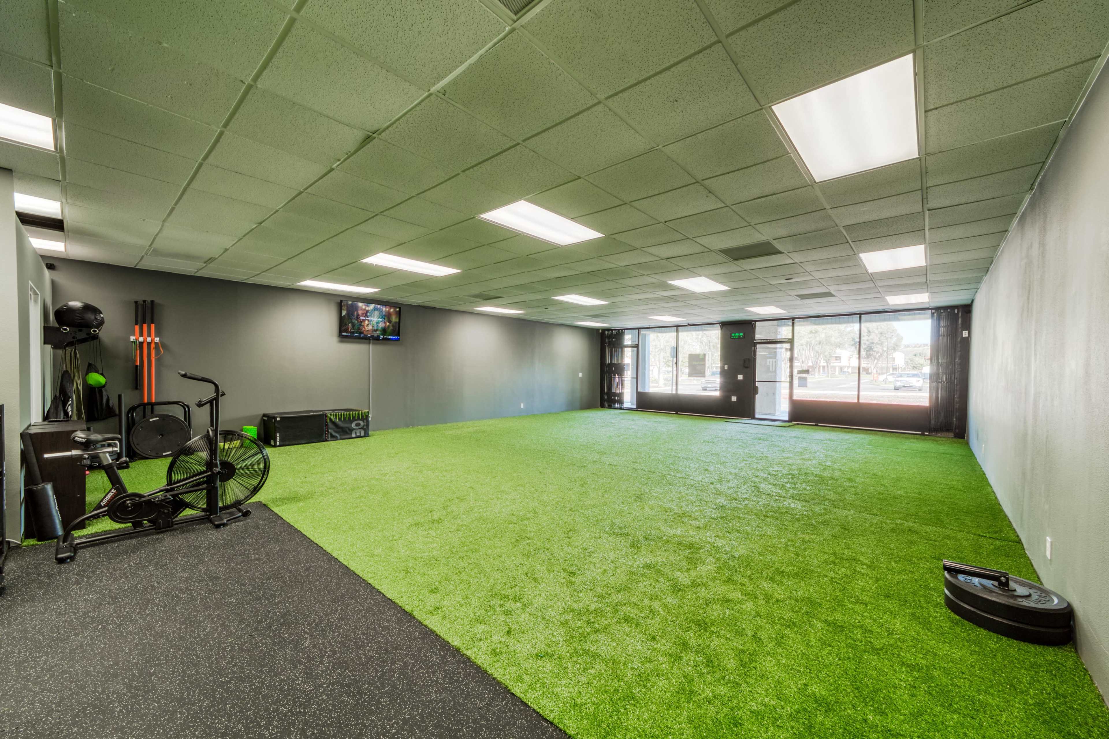 A spacious fitness studio with green turf flooring, black rubber sections, large windows, and exercise equipment along one wall.