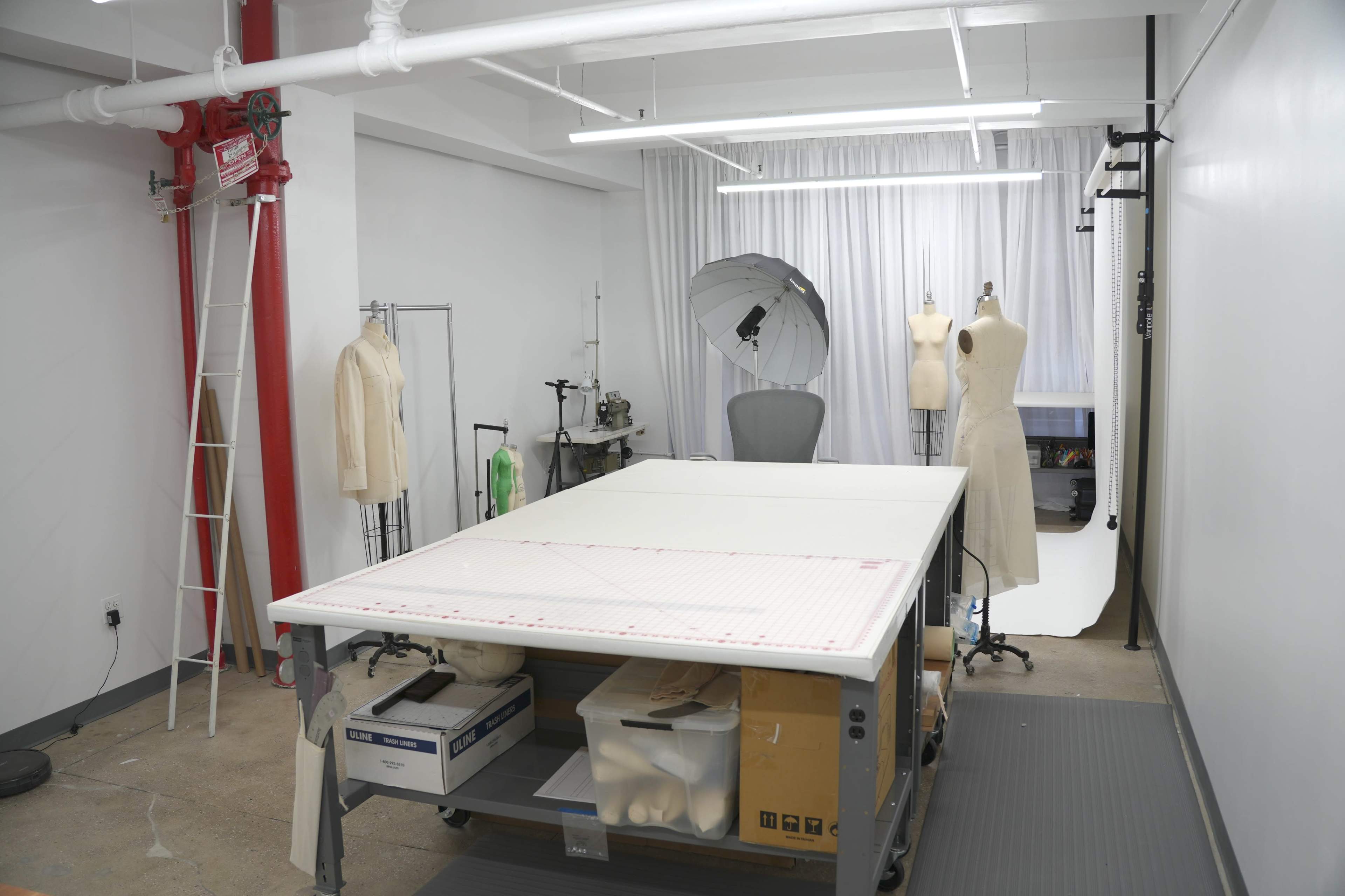 The image shows a well-equipped fashion design studio featuring a large worktable, dress forms, and various sewing equipment.