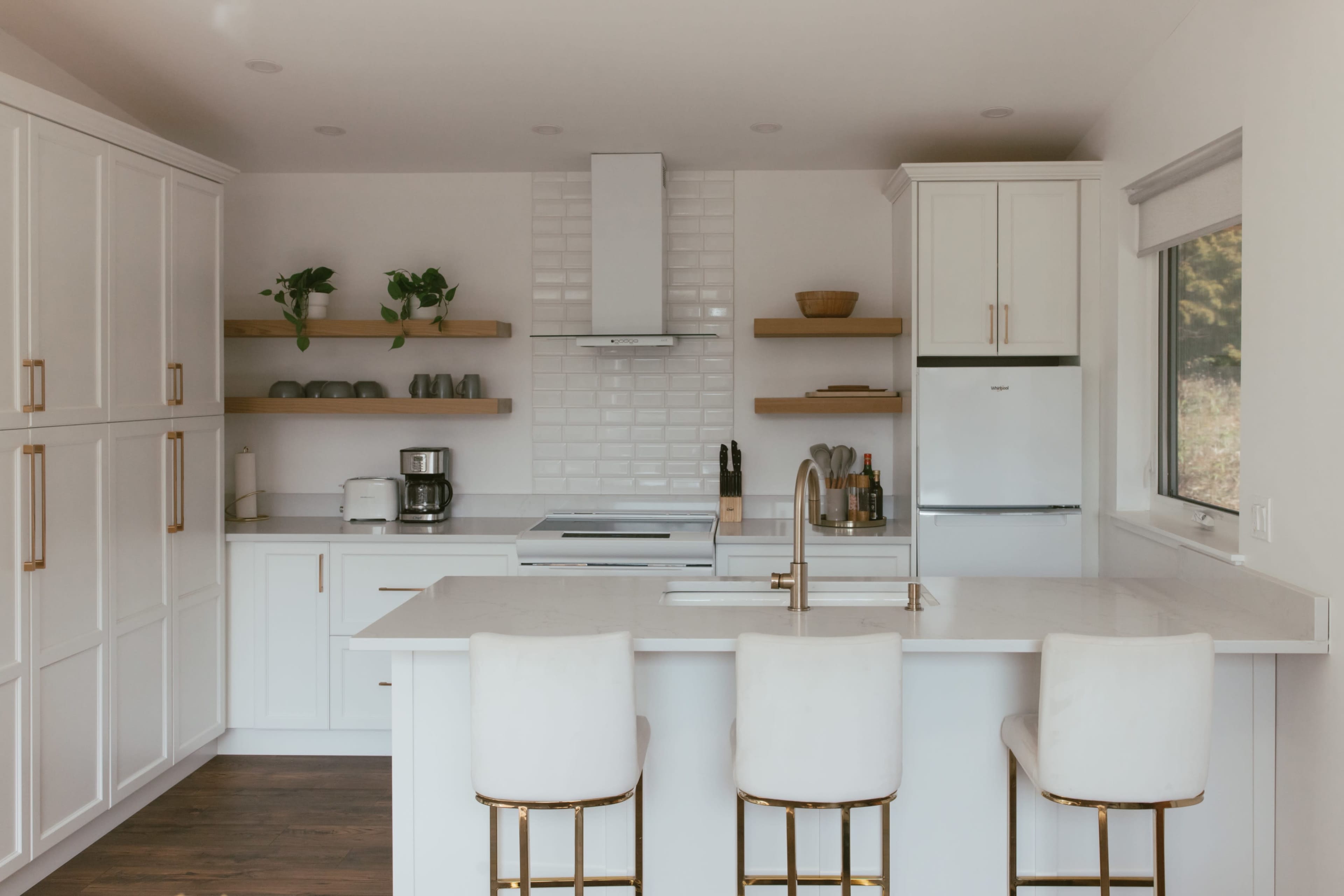 A modern kitchen features white cabinetry, a large island with bar stools, and open shelving with plants and kitchenware.