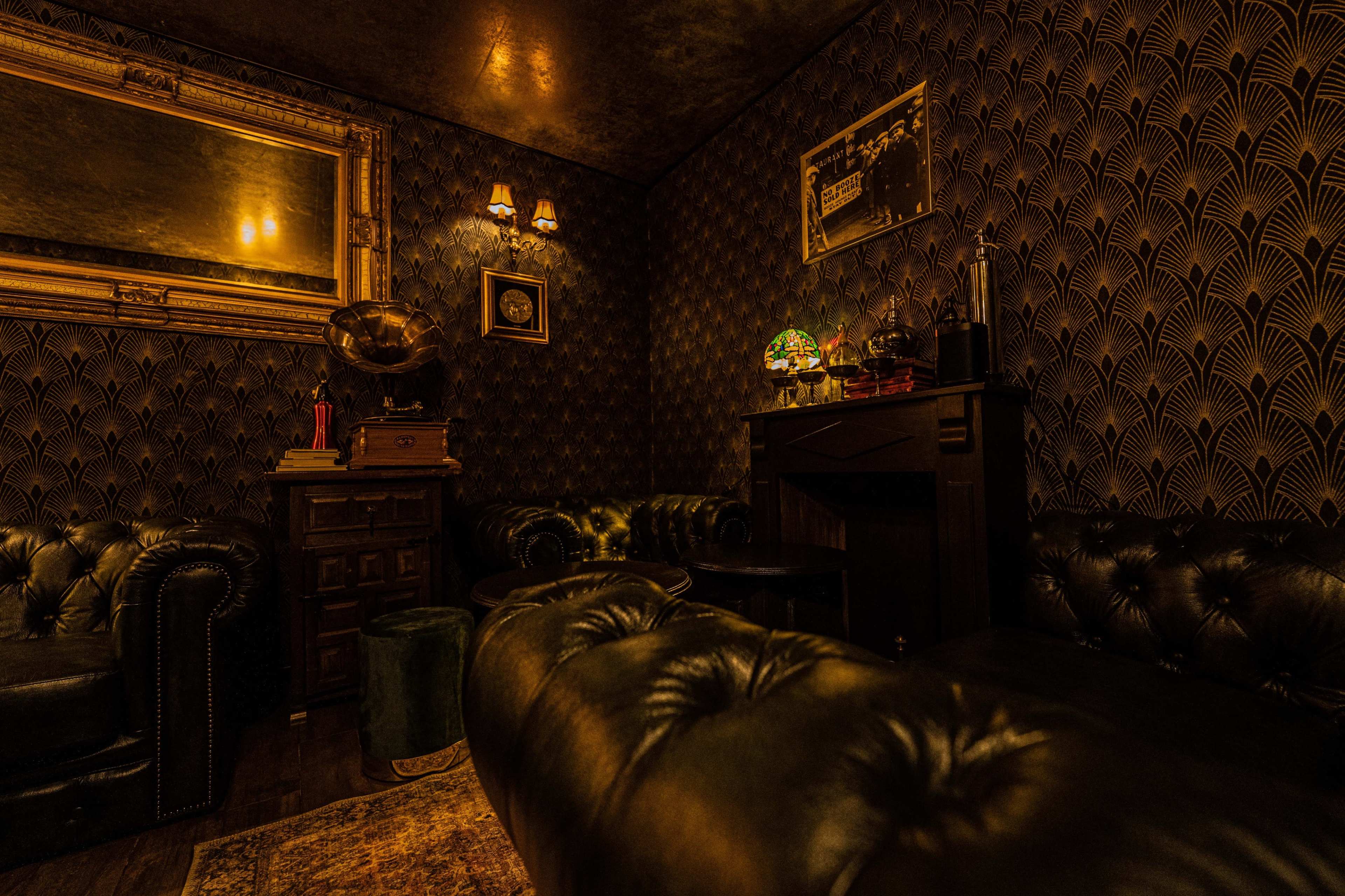 A dimly lit lounge with dark wallpaper, leather chesterfield sofas, and vintage decor, including a gramophone and a framed picture on the wall.