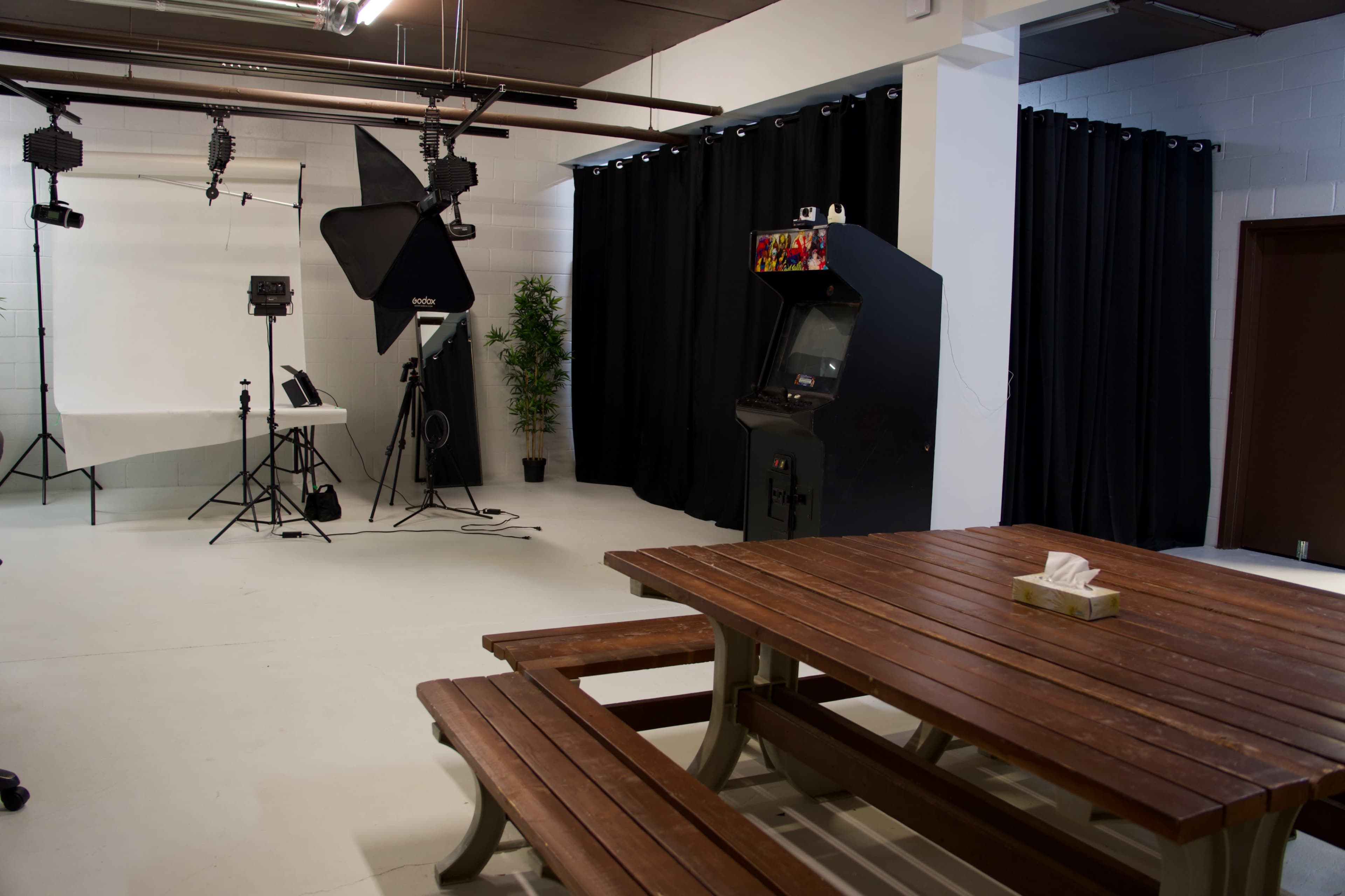 The image shows a studio space with a wooden picnic table, an arcade game machine, and photography equipment set against black curtains.