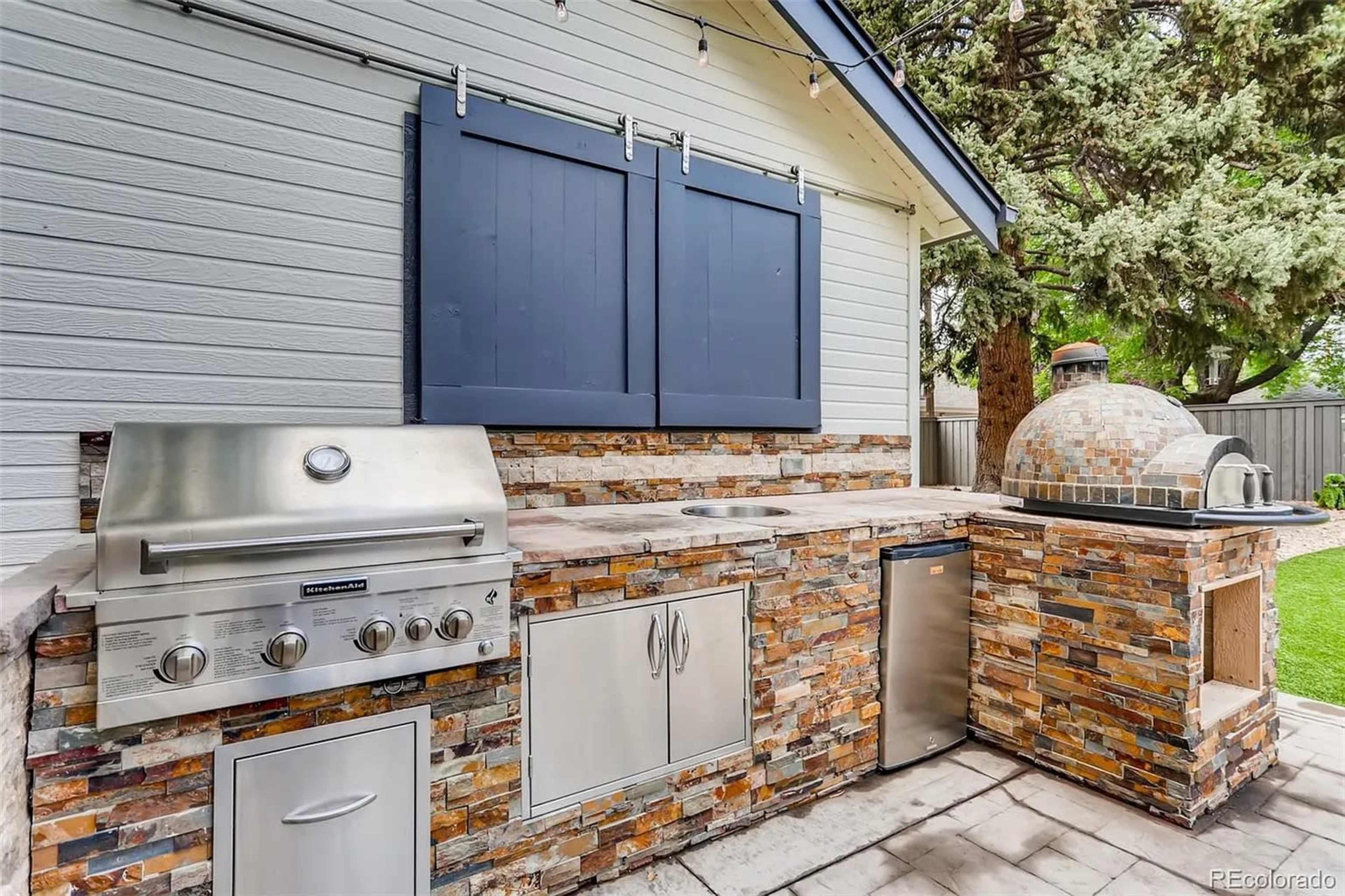 The image shows an outdoor kitchen equipped with a stainless steel grill, a stone countertop, a sink, and a pizza oven under a blue-shuttered window.