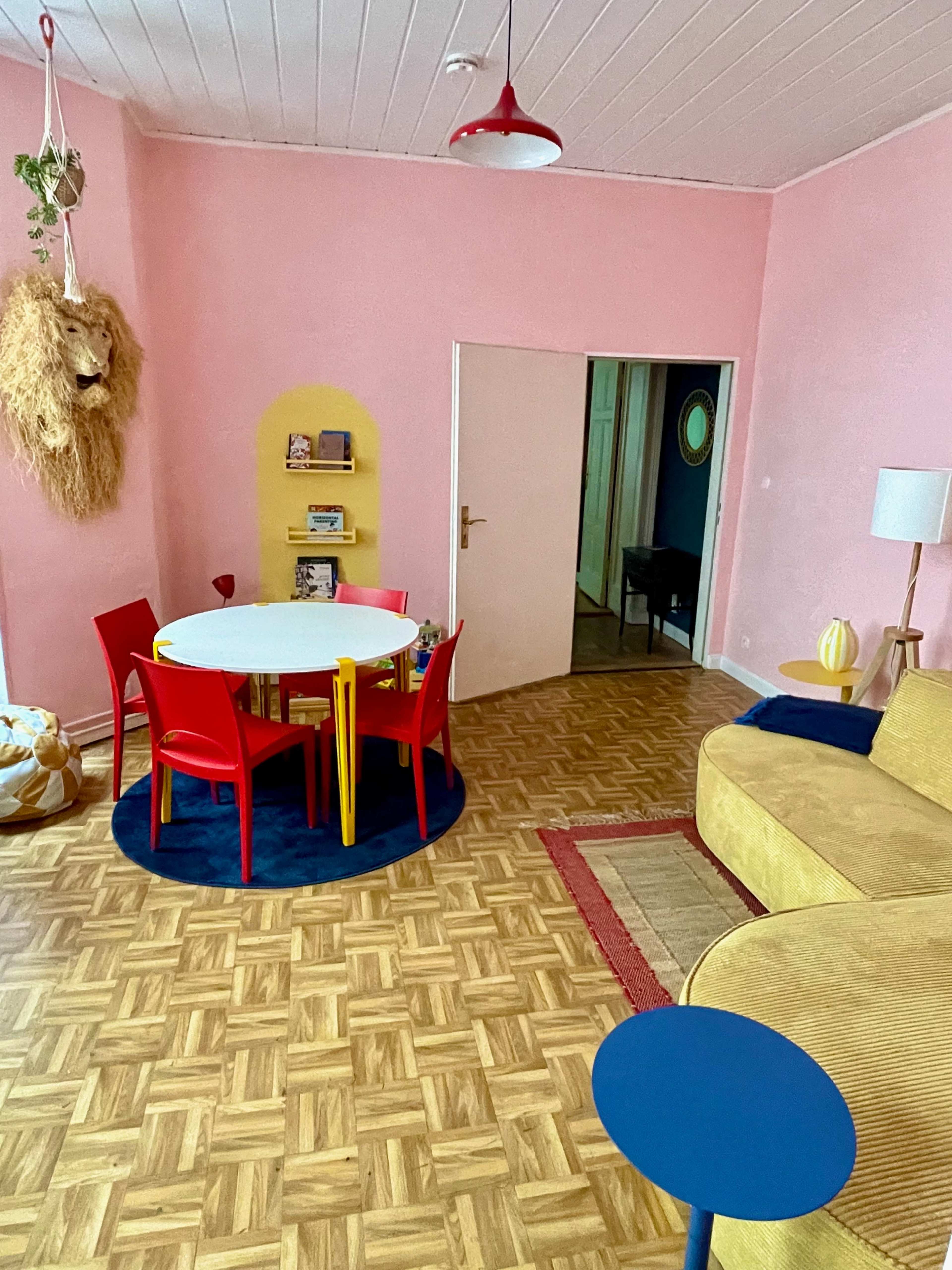 The image shows a colorful living room with a round white dining table surrounded by red and yellow chairs, a yellow accent wall, and a beige sofa.