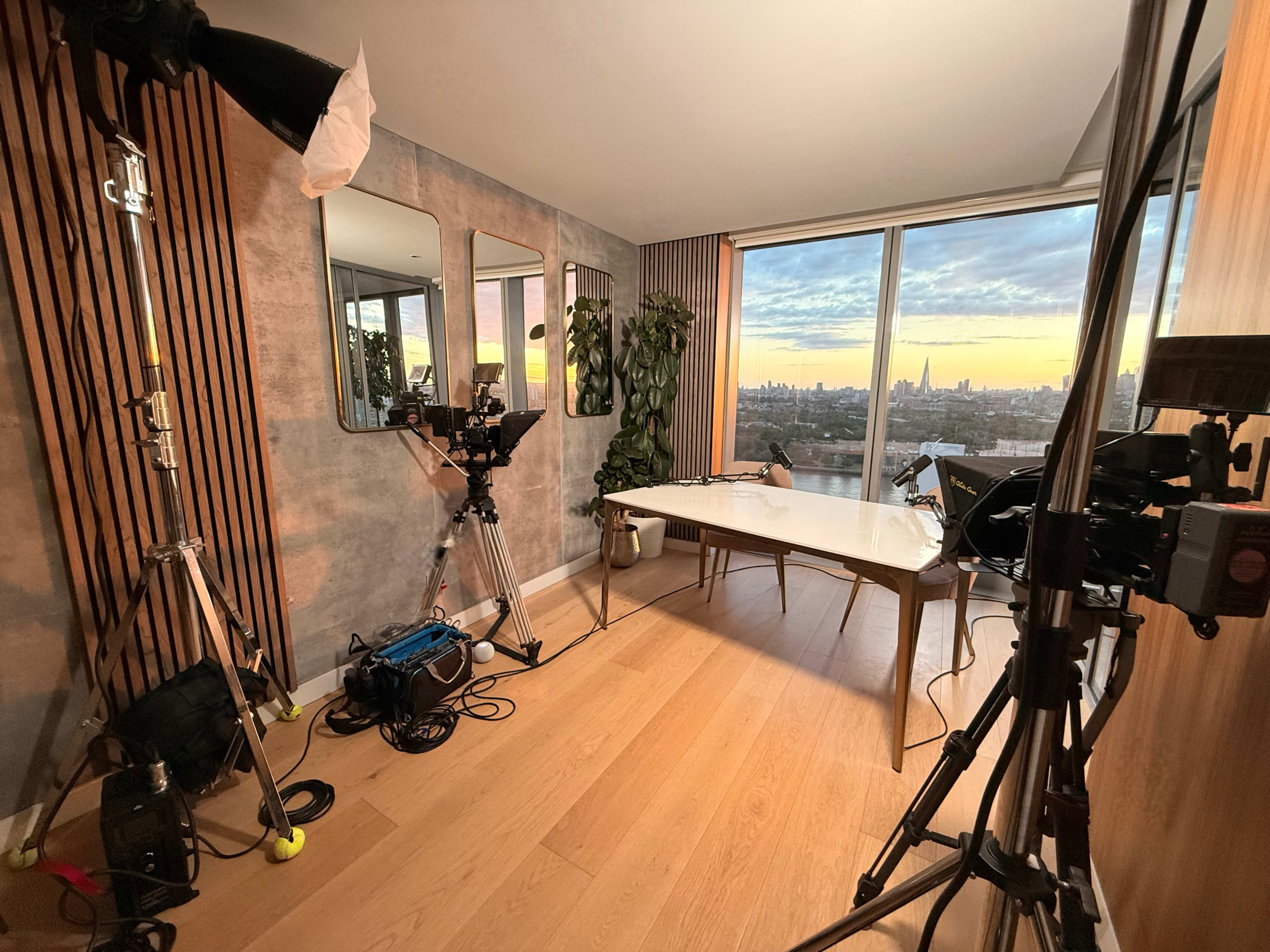 The image shows a well-lit interior studio setup with a table, camera equipment, and large windows overlooking a city skyline at sunset.