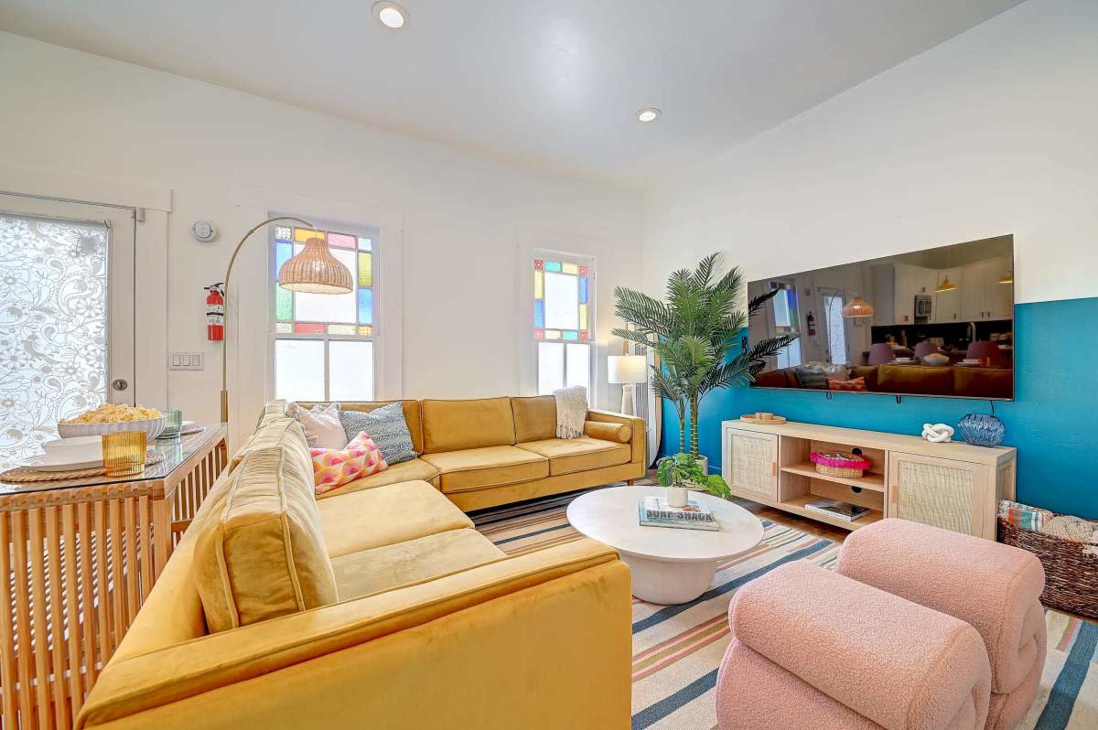 The living room features a large yellow sectional sofa, a round coffee table, and a wall-mounted television, with colorful stained glass windows and a potted plant.