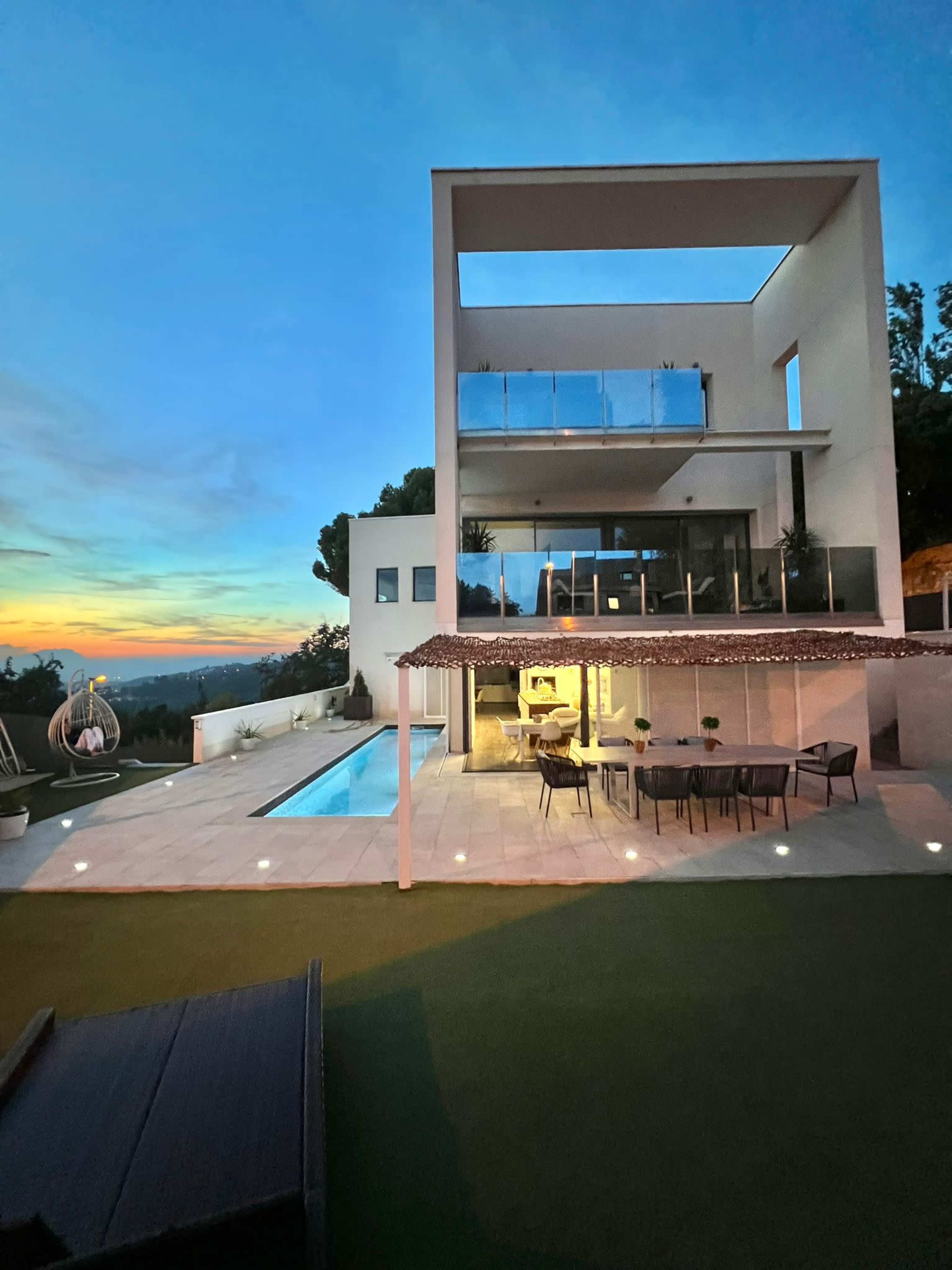 A modern house with multiple balconies overlooks a sunset, featuring a swimming pool and outdoor seating area.