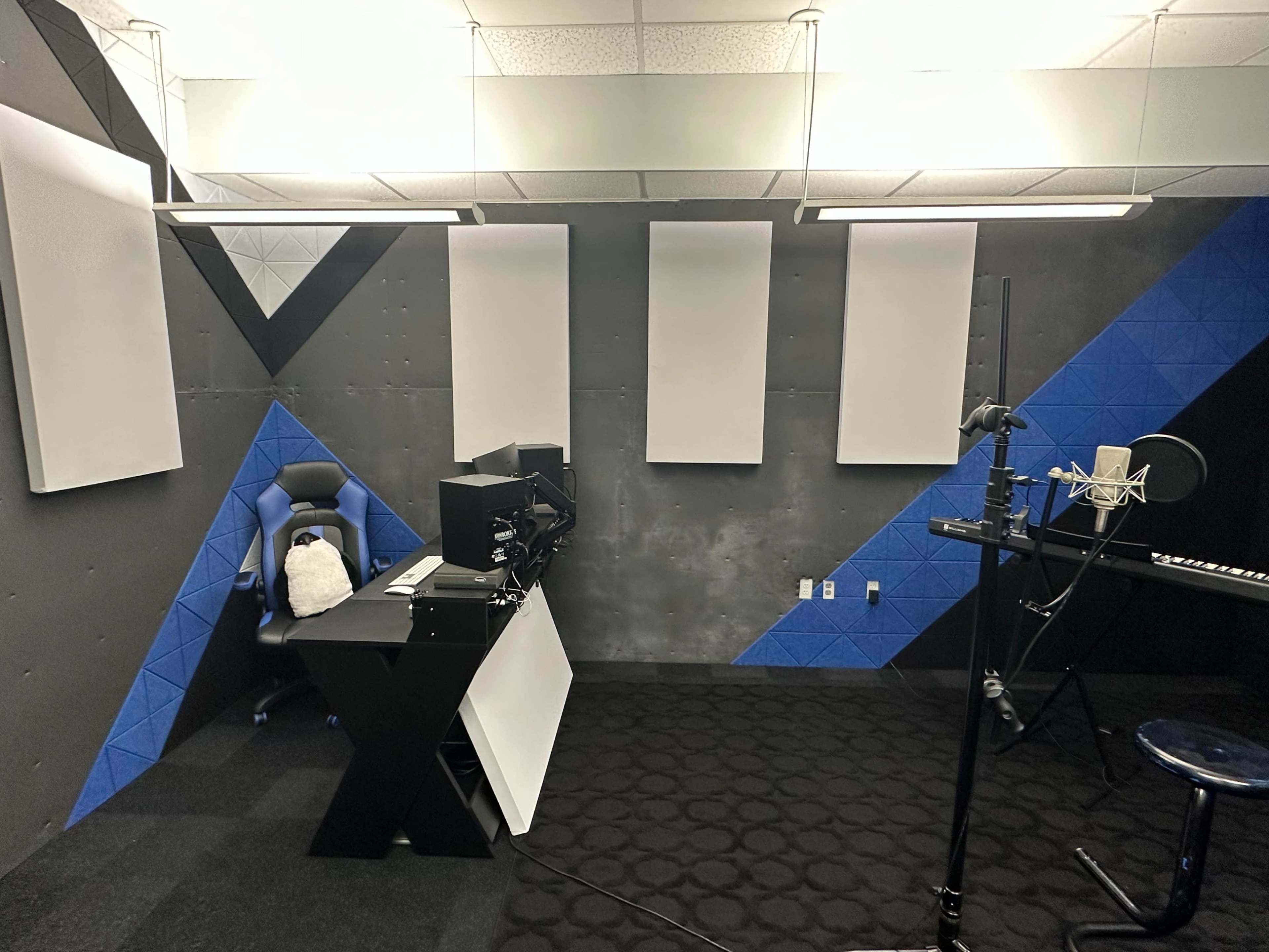 A recording studio with acoustic panels on the walls, a black and blue office chair, a desk with a computer, and a microphone setup.