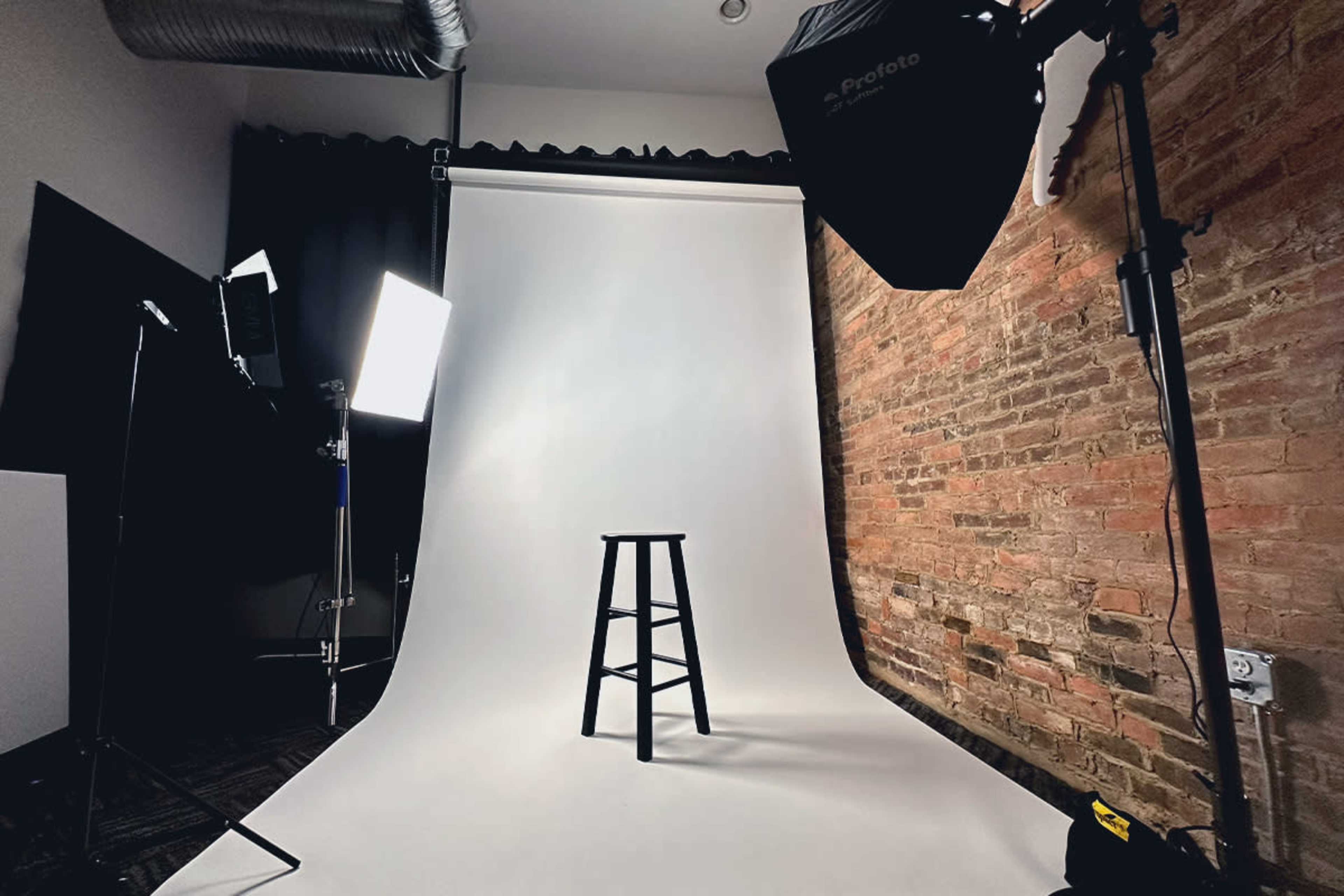 A photography studio features a black stool in front of a white backdrop, with two lights and a brick wall.