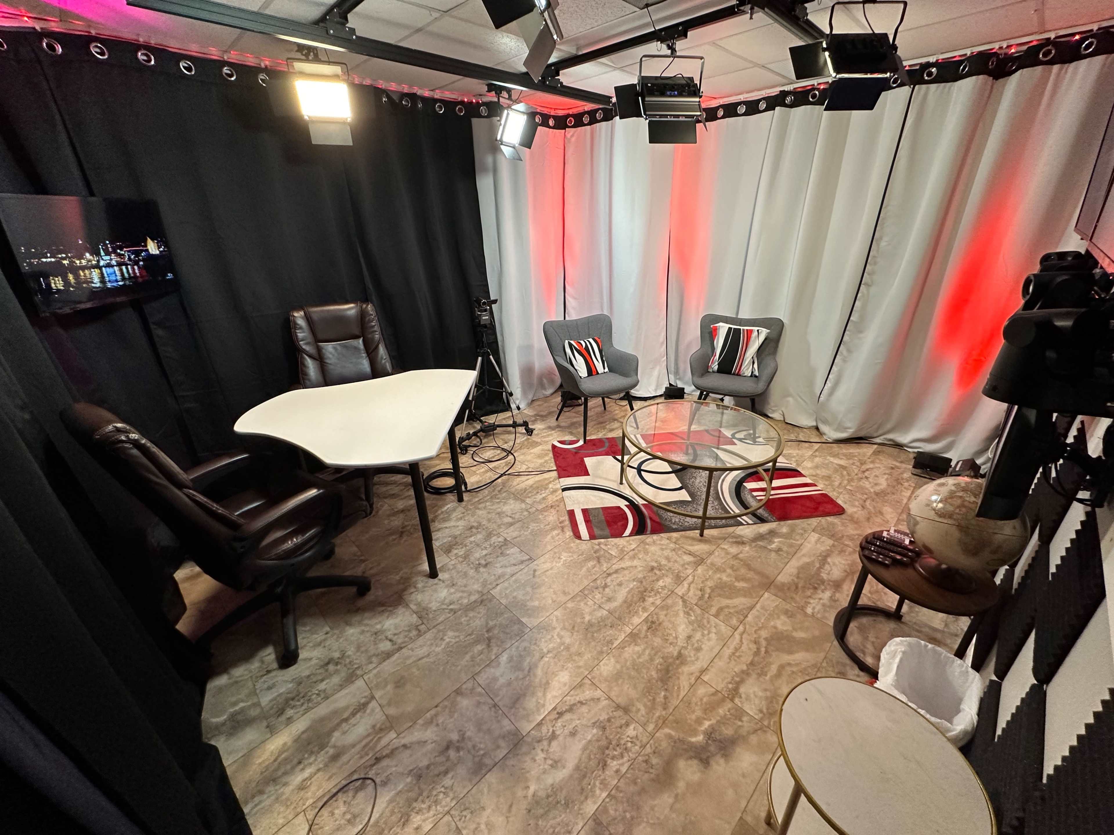 A small recording studio with two chairs, a table, a glass coffee table, and studio lights arranged on a patterned rug.